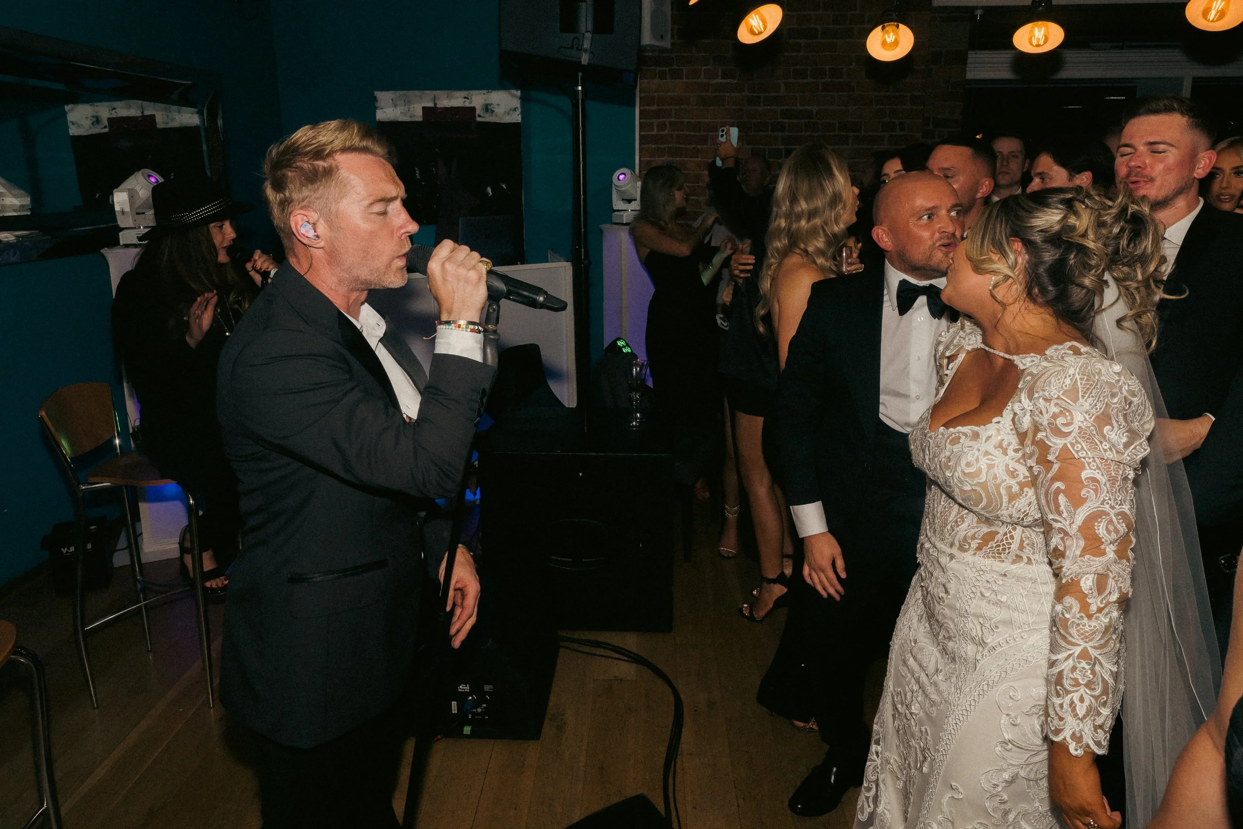 A wedding reception scene with a bride and groom kissing, a singer performing in the foreground, and guests dancing and socializing indoors with warm lighting and dark brick walls.