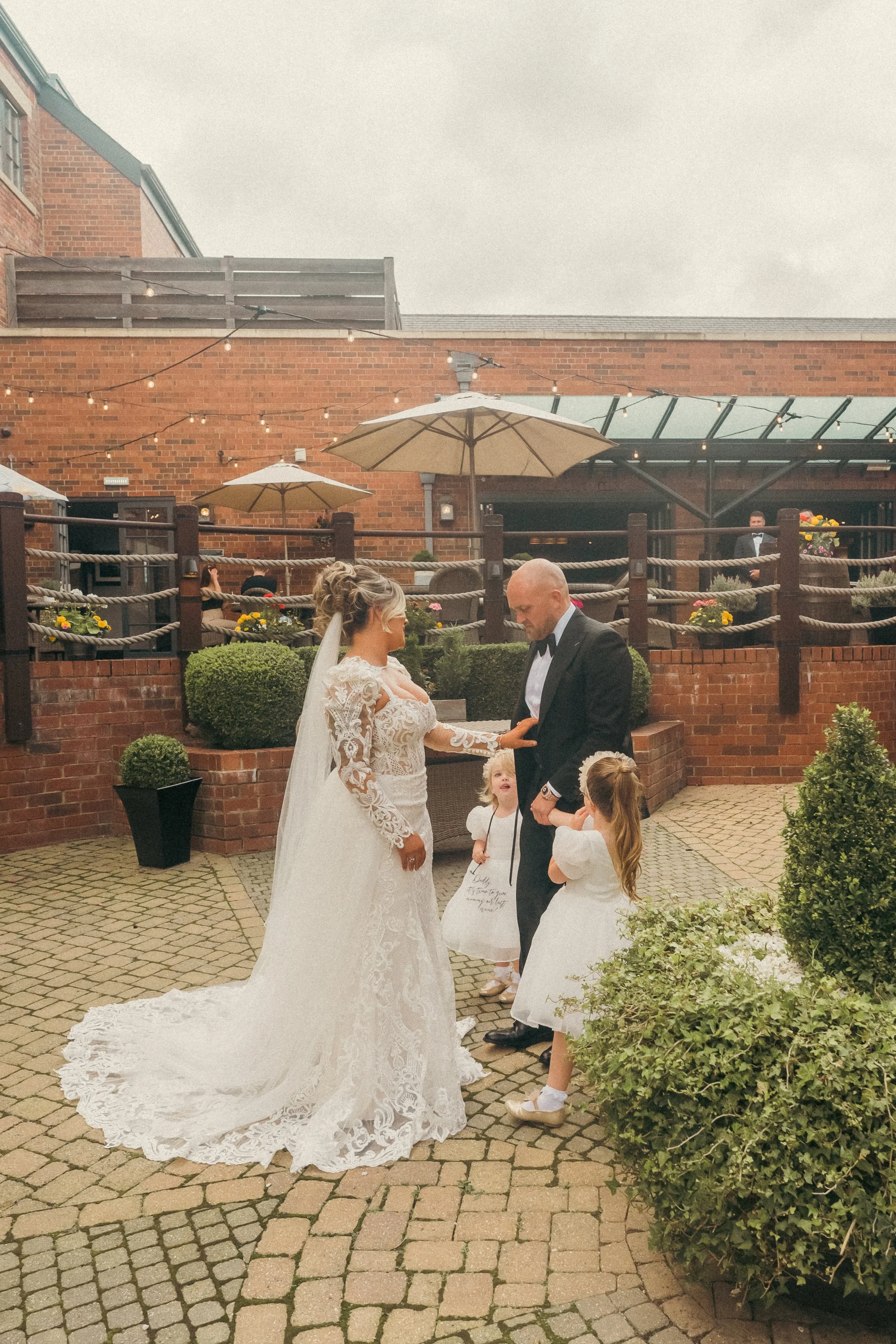 Bride and groom standing outdoors on a wedding day, surrounded by flower girls and guests, under umbrellas and string lights, in a courtyard with brick walls and greenery.