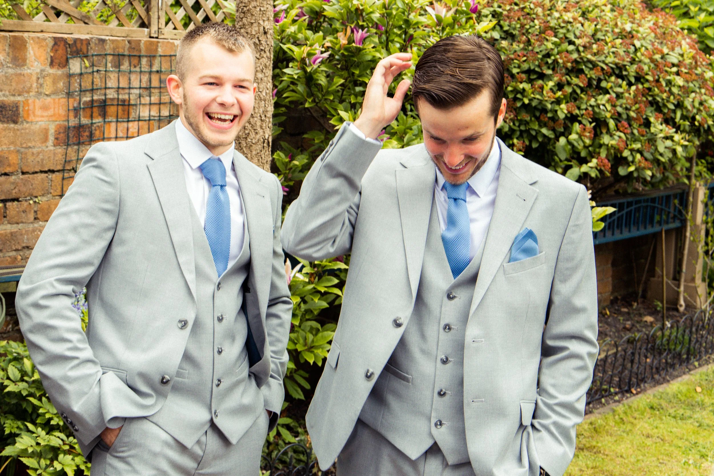 Two men in light gray suits and blue ties standing outdoors near a garden with bushes and trees, smiling and laughing, one touching his hair.