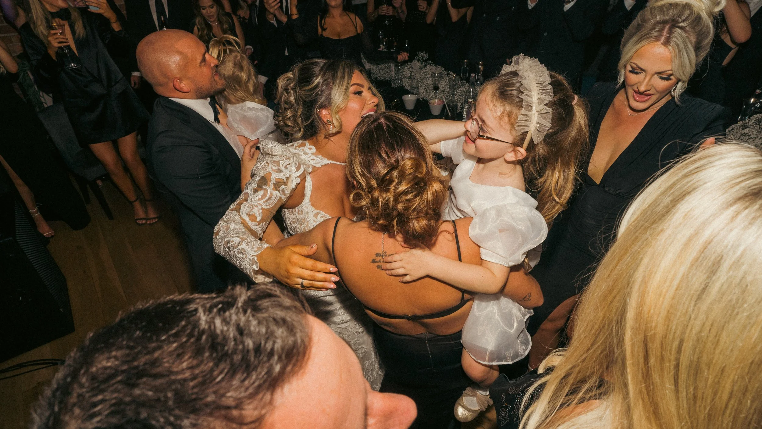 People dancing and celebrating at a party, including a woman in a white lace dress, children, and others in formal attire.