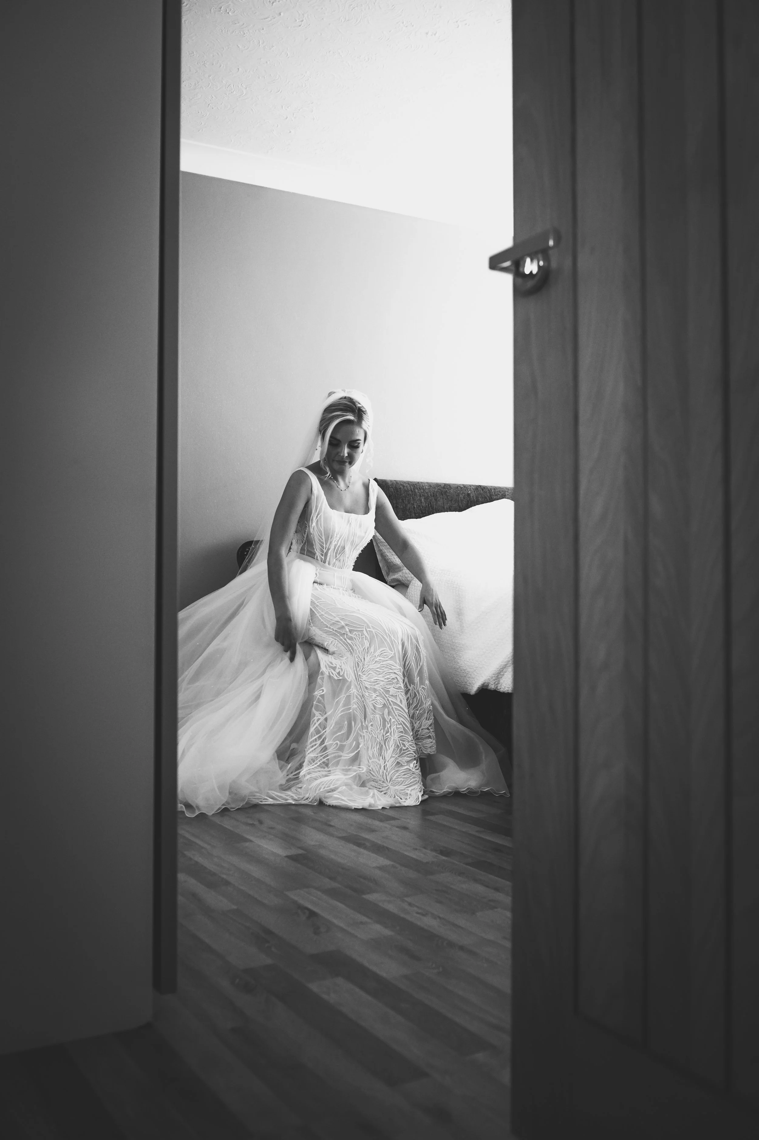 A bride in a wedding dress sitting on a bed, viewed through a doorway in black and white.