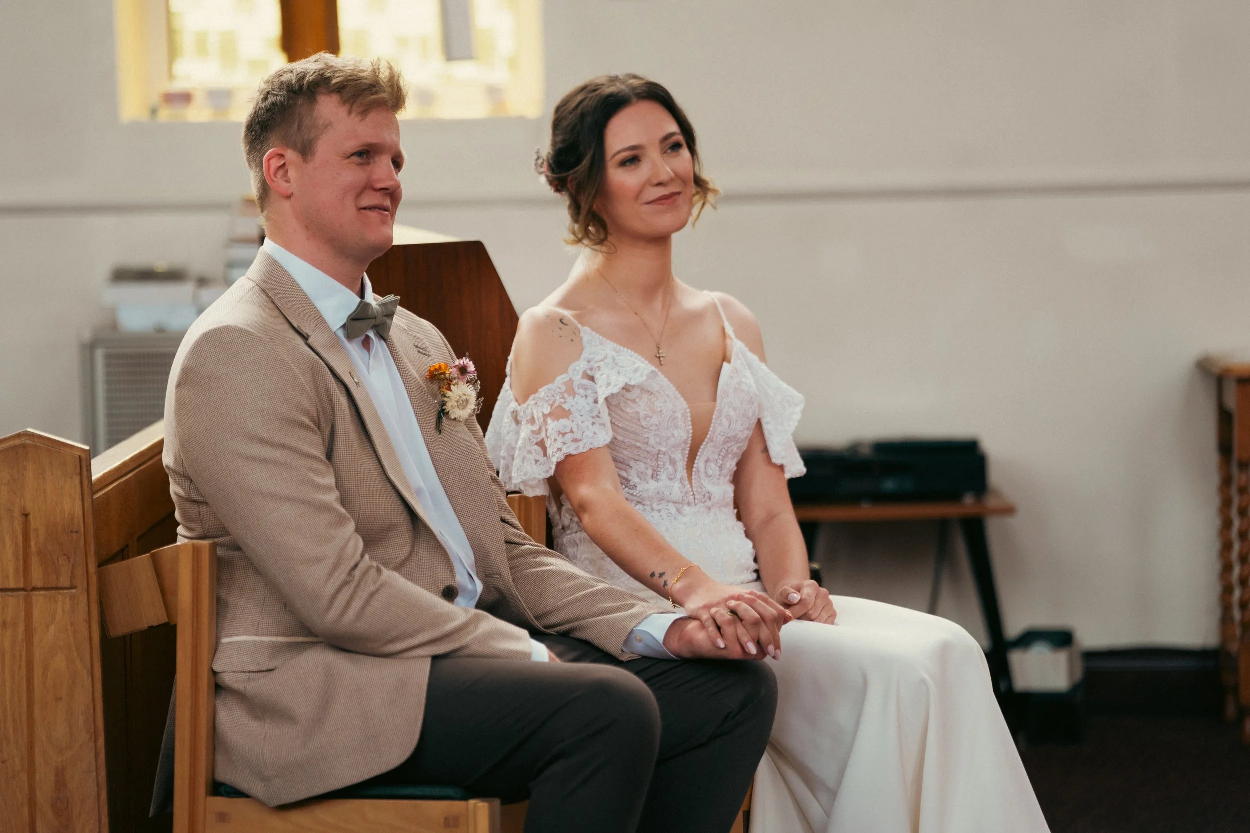 A bride and groom sit hand in hand during a wedding ceremony in a church, smiling softly. The groom wears a beige suit with a bowtie, and the bride wears a white lace dress with cold shoulder sleeves and a cross necklace.