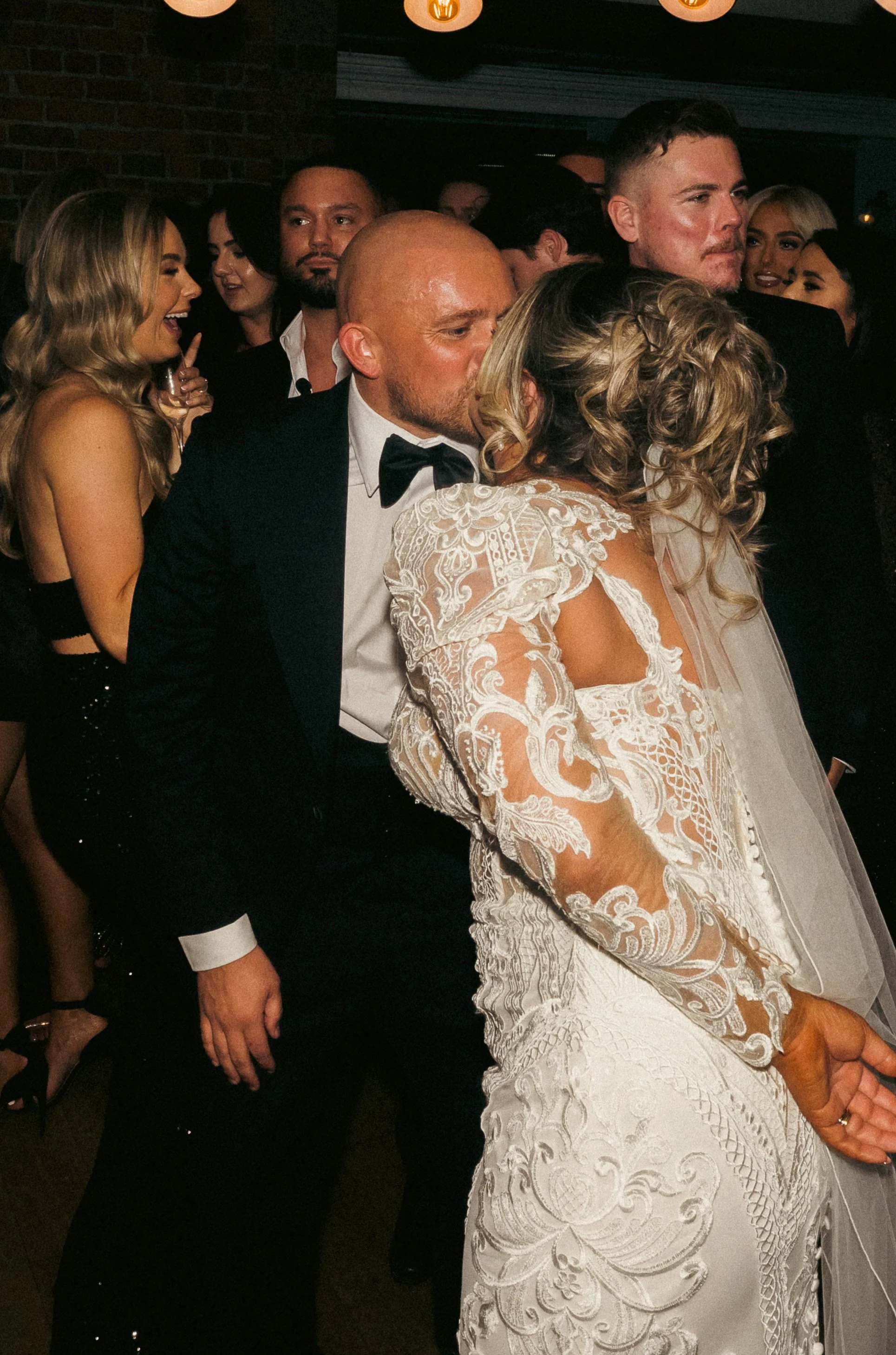 A group of people at a wedding, with a bride and groom sharing a kiss in the foreground, surrounded by guests, some of whom are smiling and laughing.