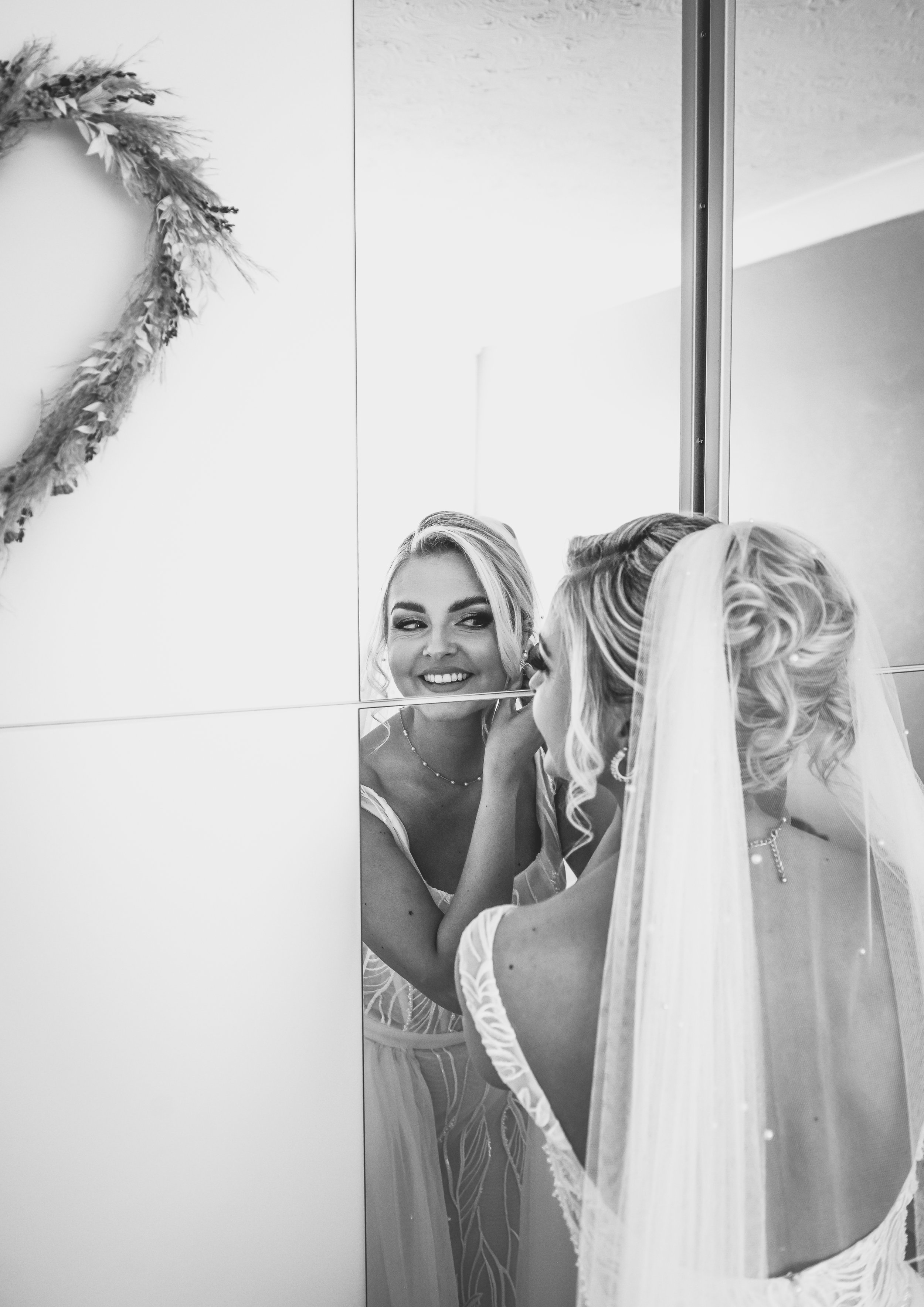 A bride smiling as she gets her makeup done in front of a mirror, reflected in a large mirror with a floral wreath decoration on the wall.