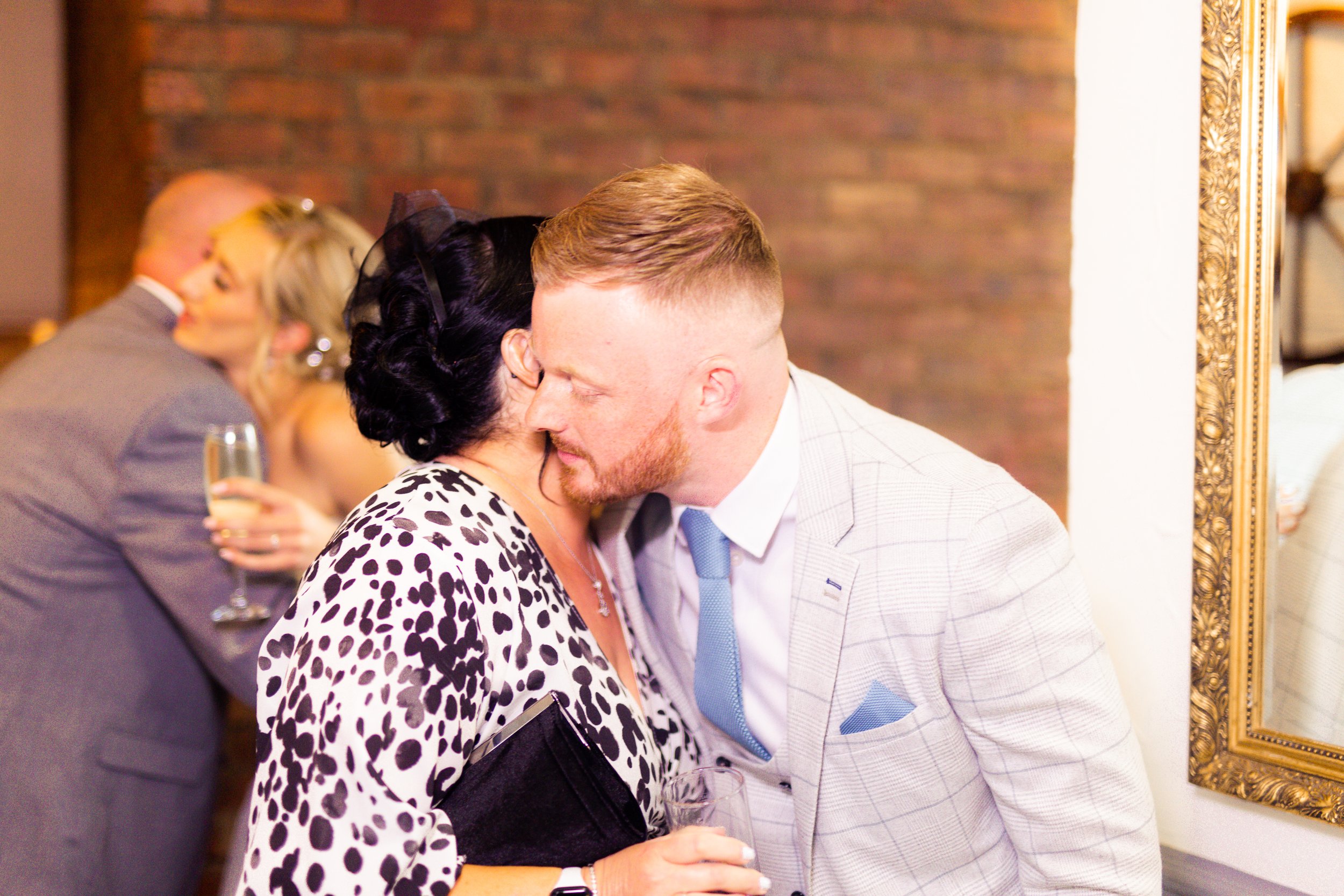 A man and woman are sharing a close moment, leaning in for a kiss or whispering at a social event. The woman has dark hair styled in an updo and is holding a glass of champagne, wearing a black and white animal print dress. The man has short, light h
