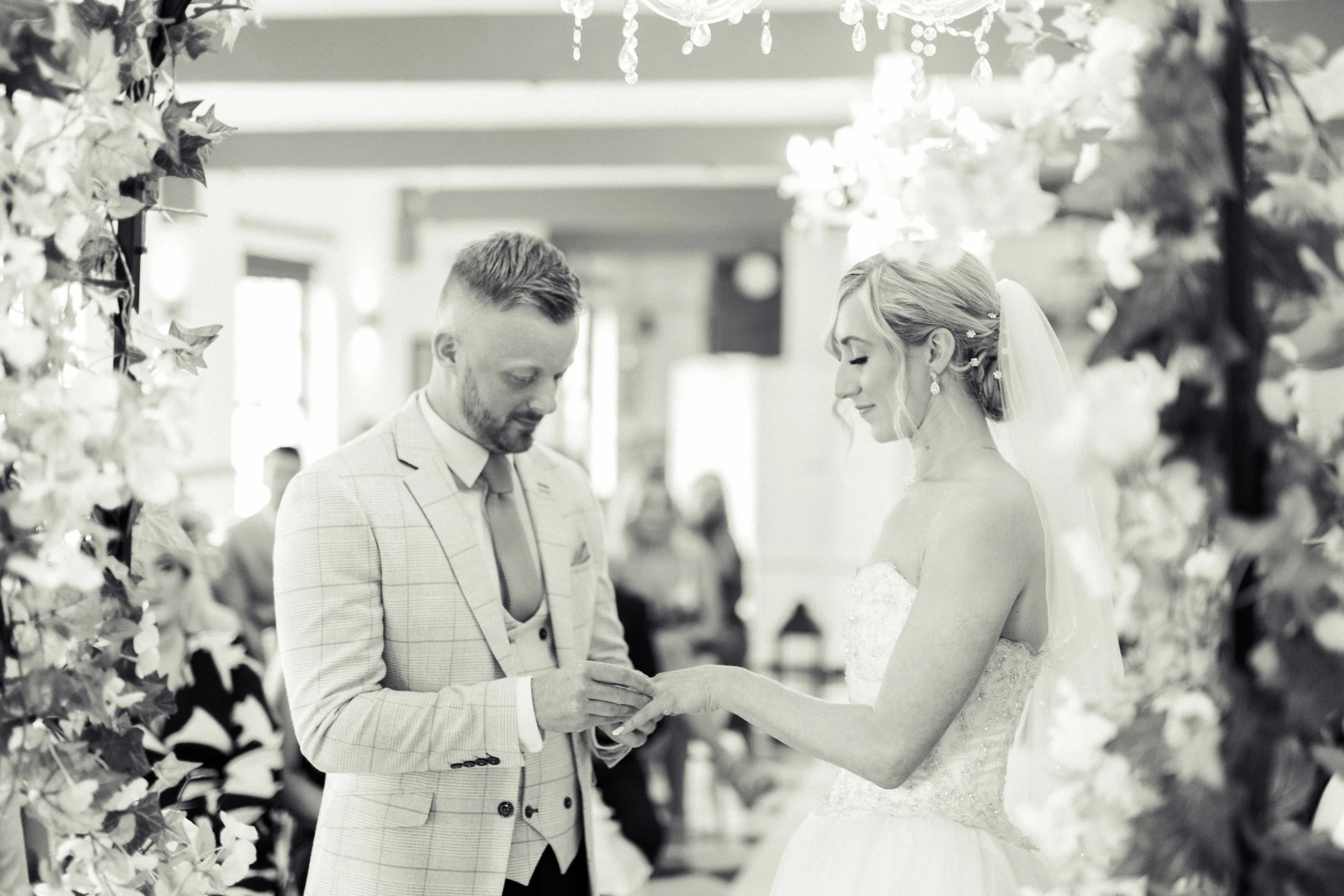 Black and white photo of a bride and groom exchanging rings during wedding ceremony surrounded by floral decorations.