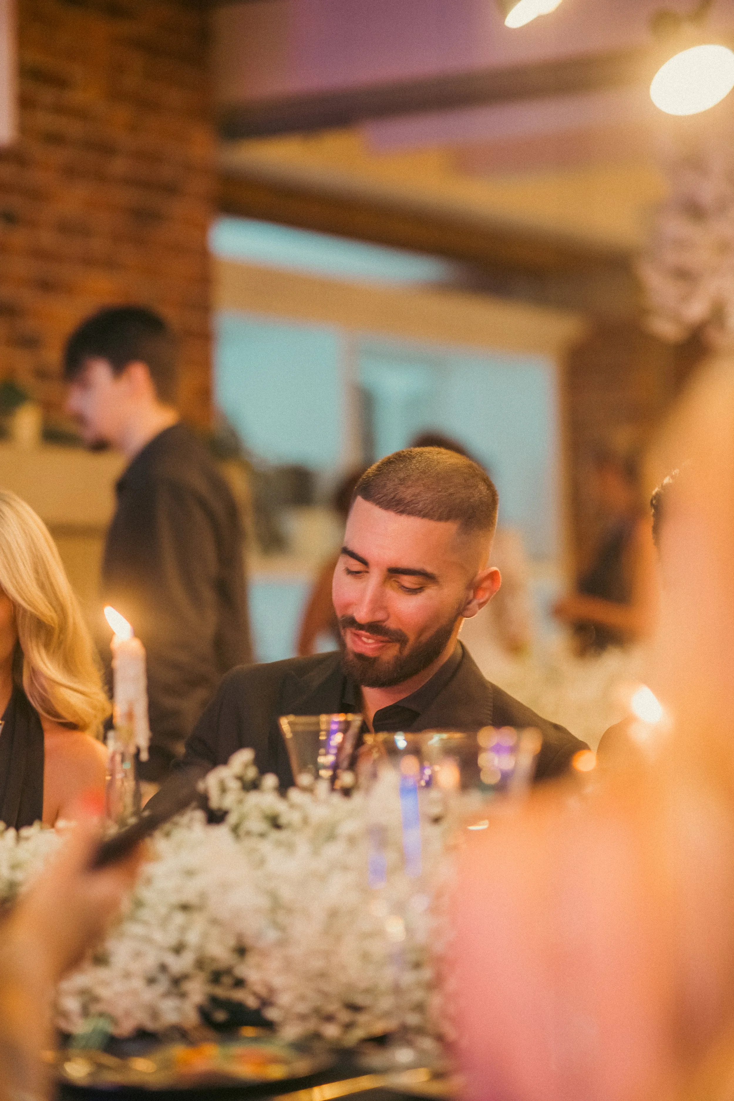 Man with a beard and short haircut at a dinner table, smiling and looking down, decorated with candles and flowers, at a warm, cozy indoor event.
