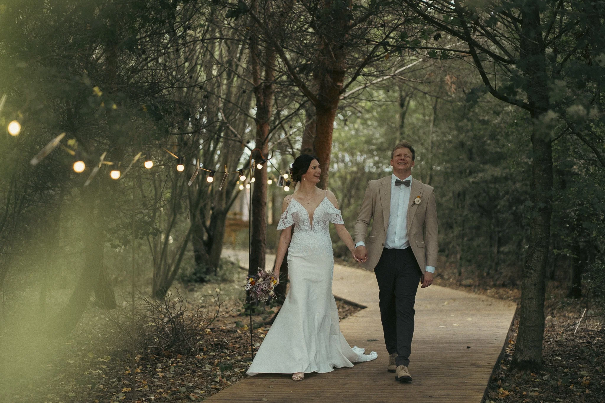 A bride and groom walking hand in hand on a forest trail lined with string lights, surrounded by trees, during a wedding celebration.