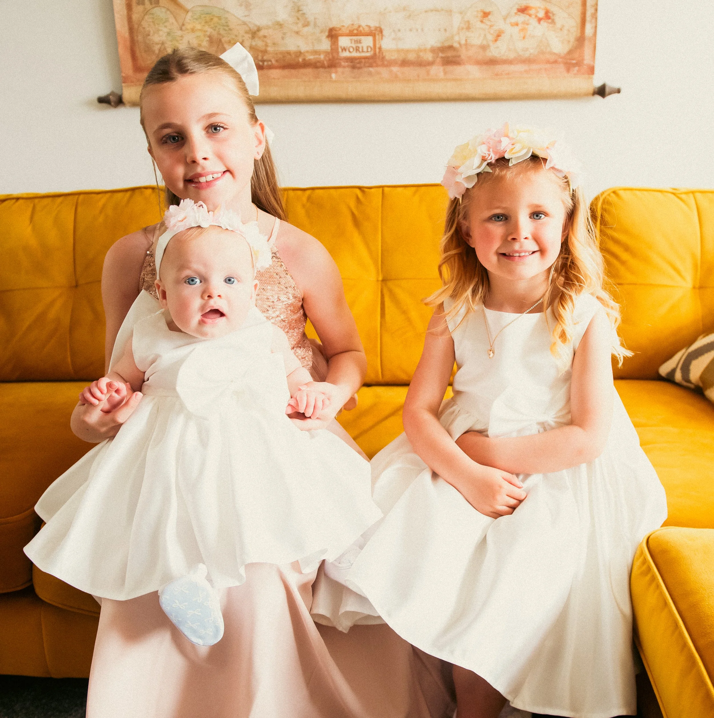 Three young girls dressed in white dresses sitting on a yellow sofa, with a world map in the background.