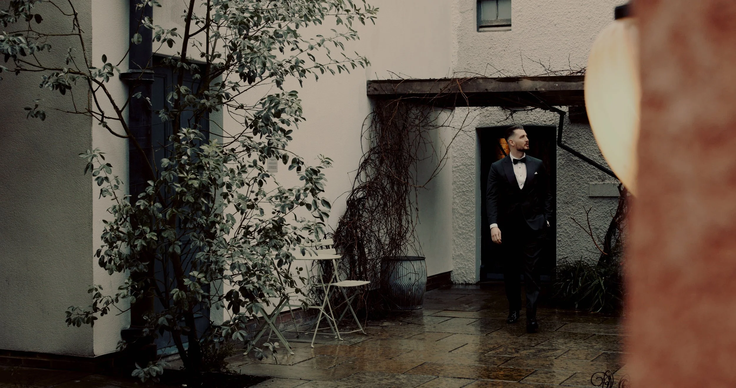 A man in a tuxedo standing outdoors near a door, with a small table and chairs on the wet ground, and a large plant on the left side of the image.