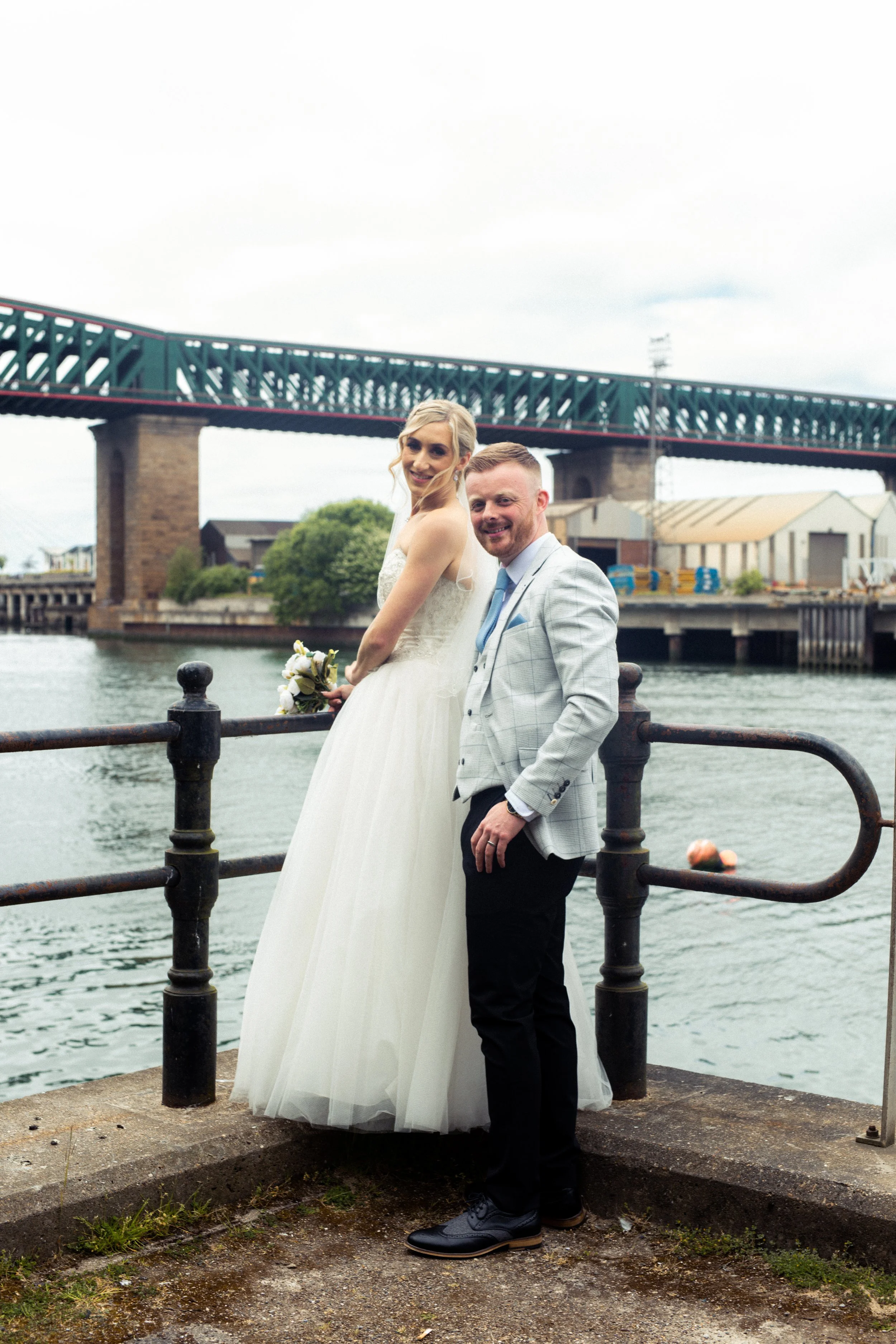 A bride in a white wedding dress holding a bouquet standing next to a groom in a light gray suit jacket, near a body of water with a bridge in the background.