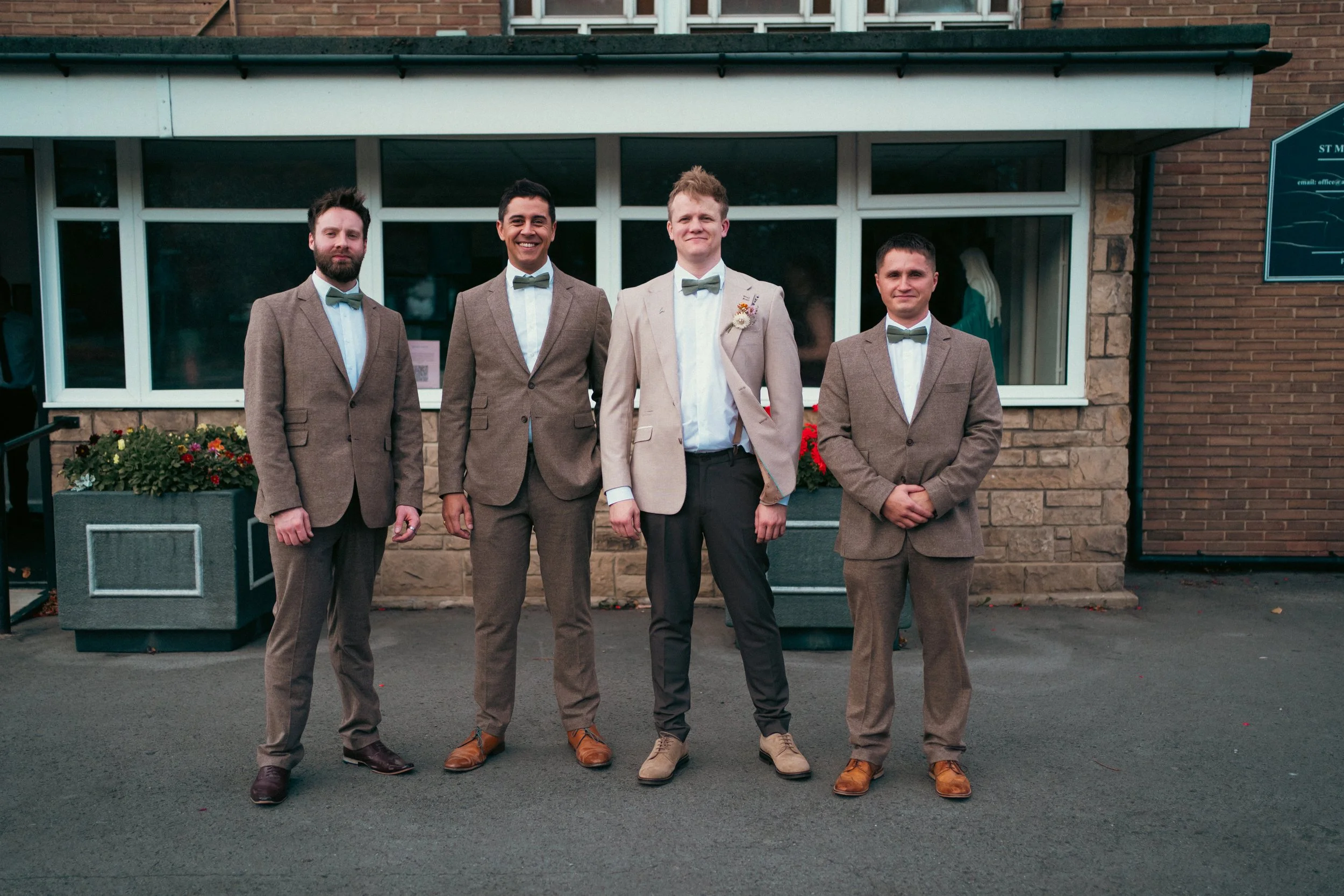 Four men standing outdoors in front of a brick building with large windows, dressed in suits with bow ties, posing for a group photo.