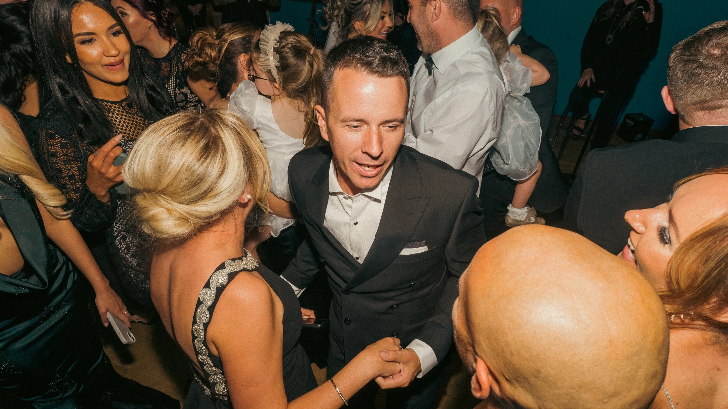 People dancing and socializing at a lively party or celebration, with a man in a tuxedo holding hands and engaging with a woman with blonde hair.
