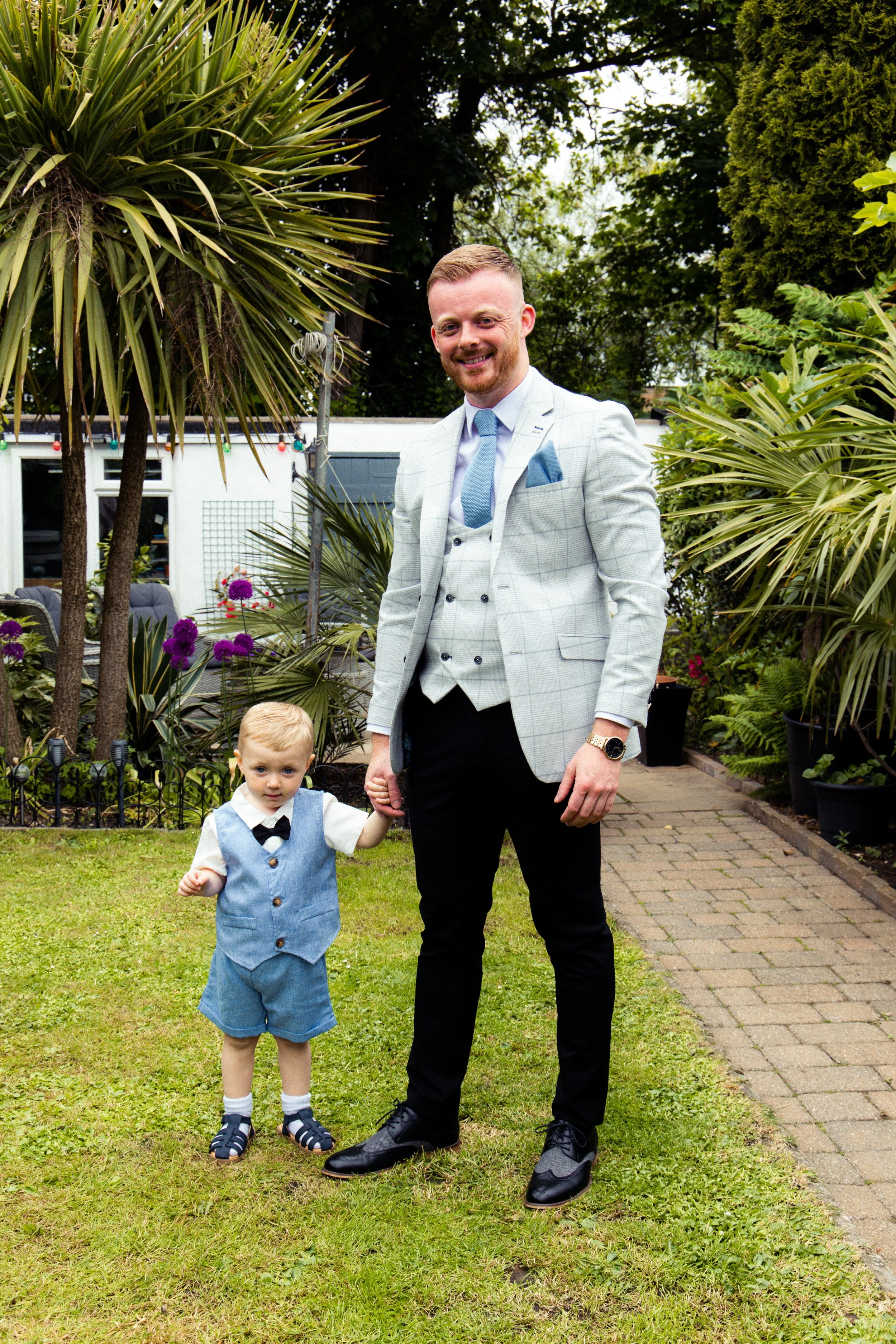 A man in a light gray plaid blazer holding hands with a young boy in a blue vest and shorts outdoors in a garden.
