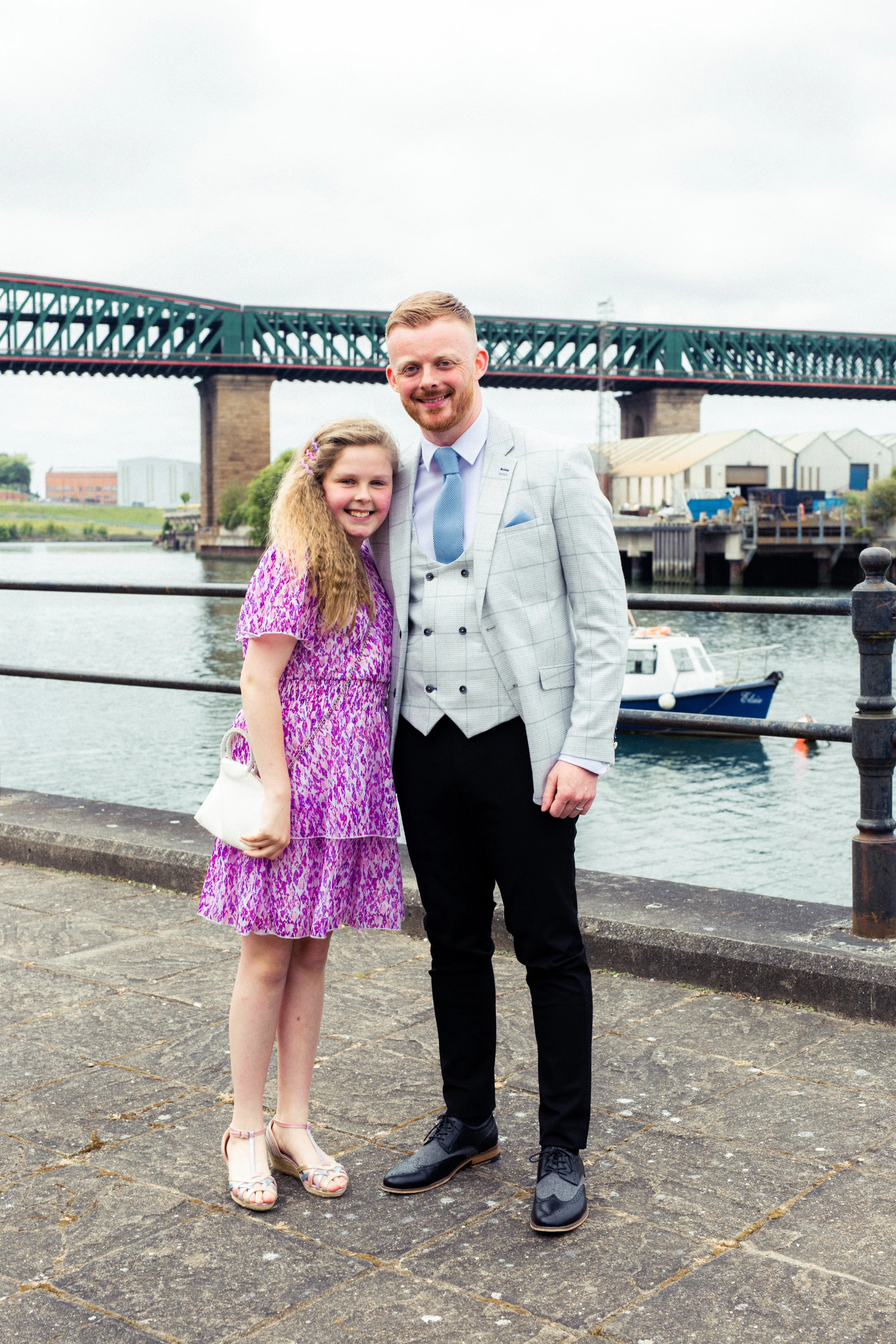 A man and a girl standing outdoors near a river, smiling at the camera with a bridge in the background.