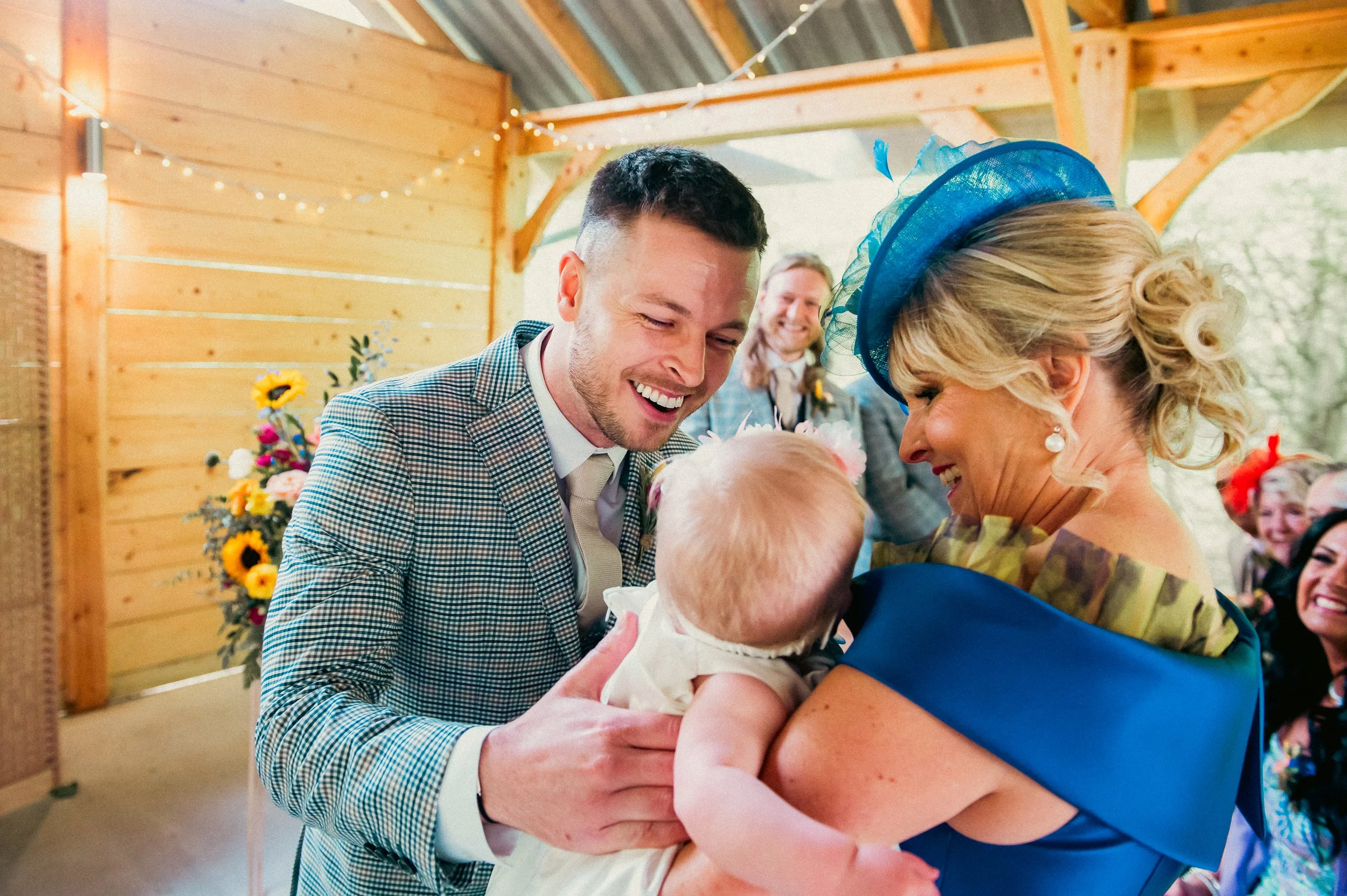 A joyful young man, woman in a blue dress with a matching hat holding a baby, with a woman in the background smiling, in a rustic wooden venue decorated with flowers and string lights.