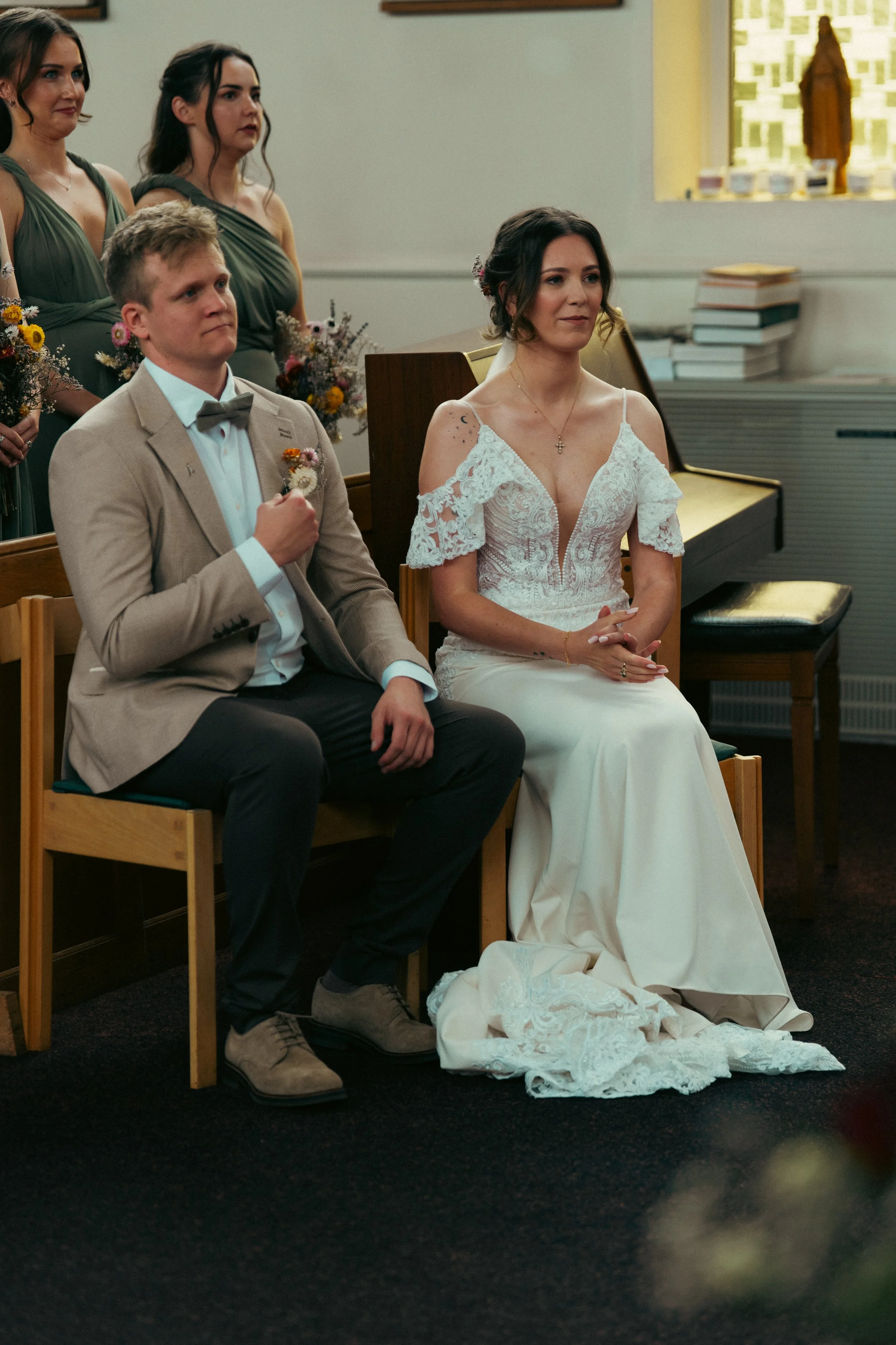 A bride and groom sitting during a wedding ceremony, with bridesmaids standing behind them in matching dresses, inside a church or chapel.