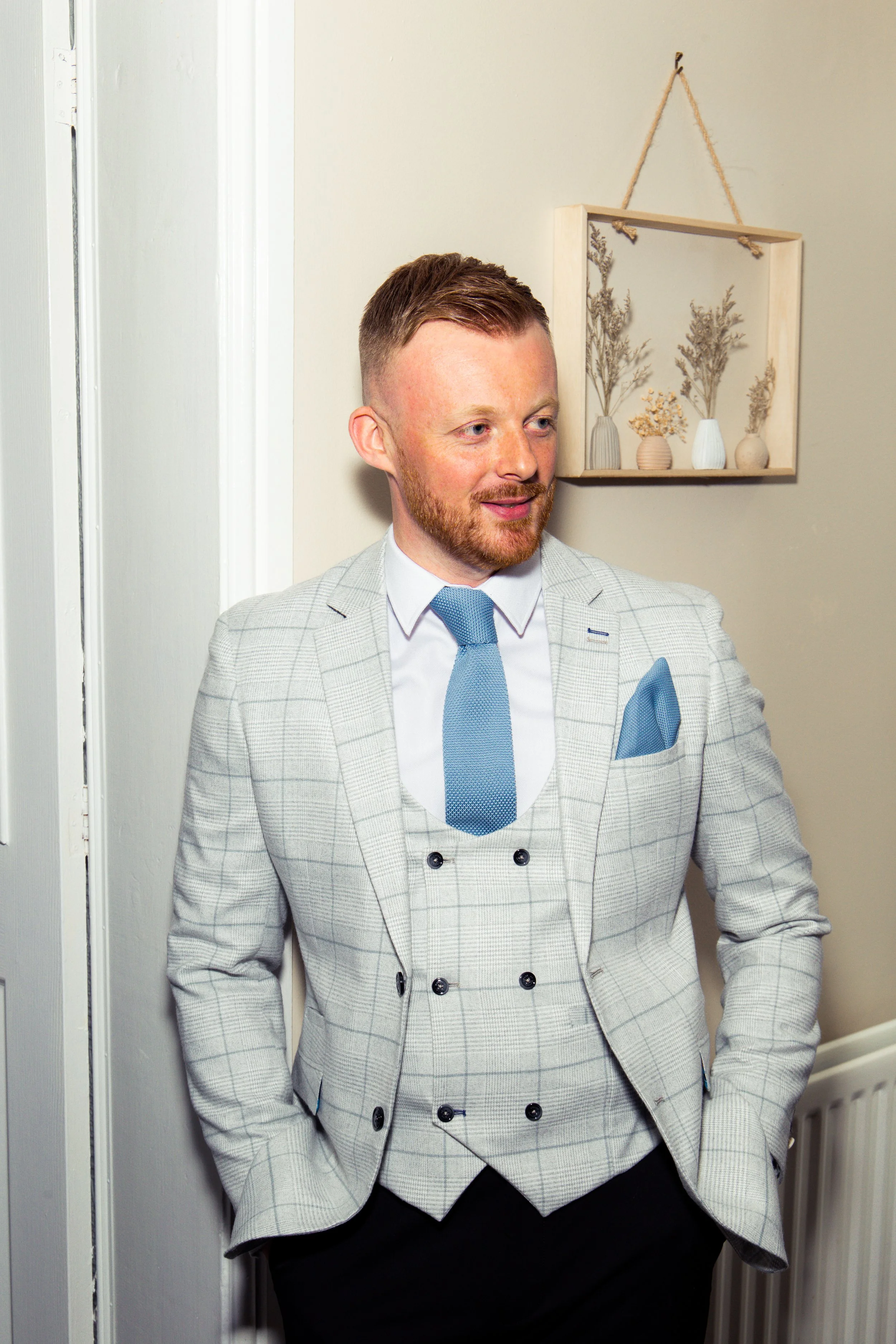 A man in a light-colored plaid suit with a blue tie and pocket square, standing indoors with his hands in his pockets, smiling slightly, with a beige wall and decorative wall hanging behind him.