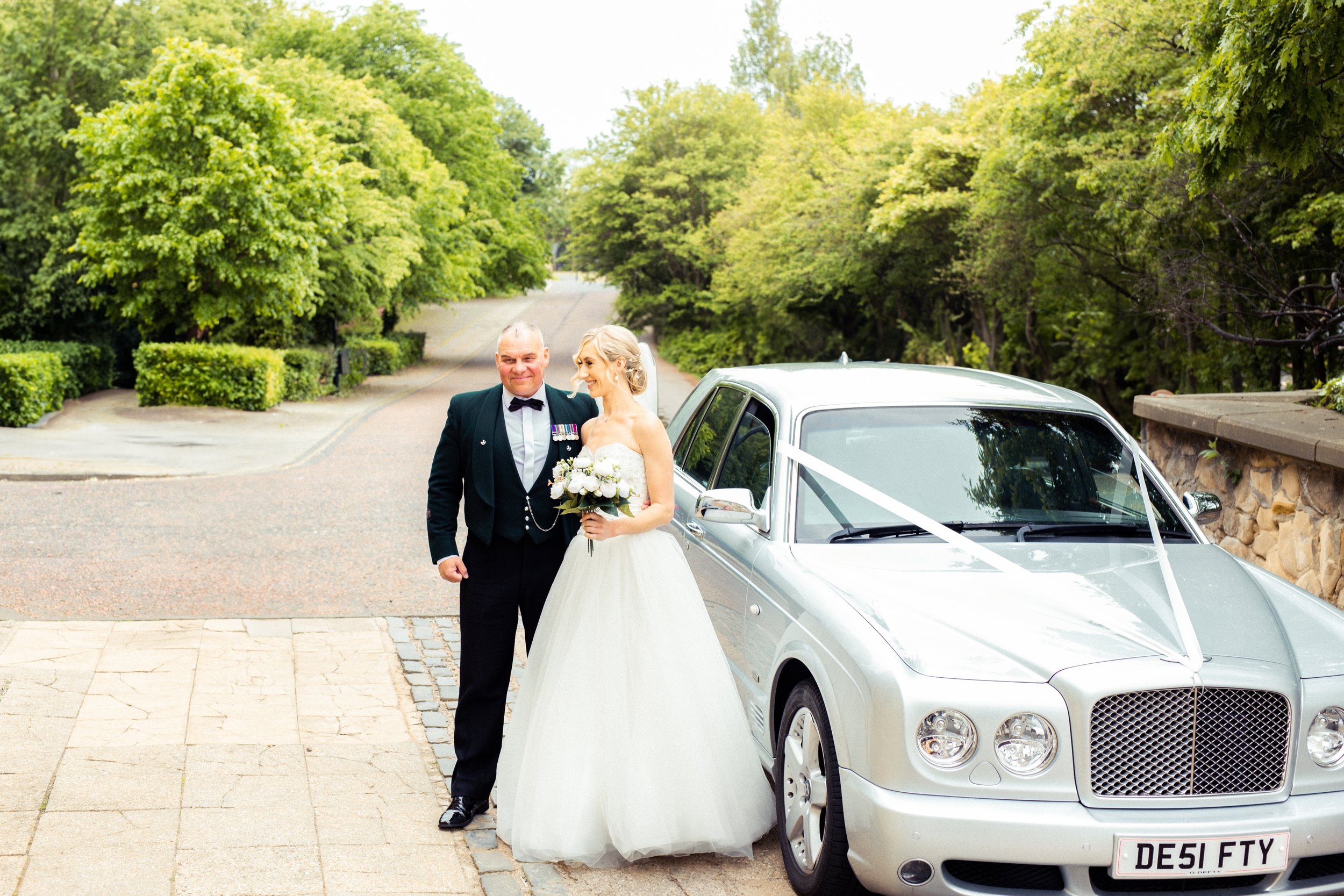 Bride and groom standing next to a decorated wedding car in a scenic outdoor setting with trees and greenery.