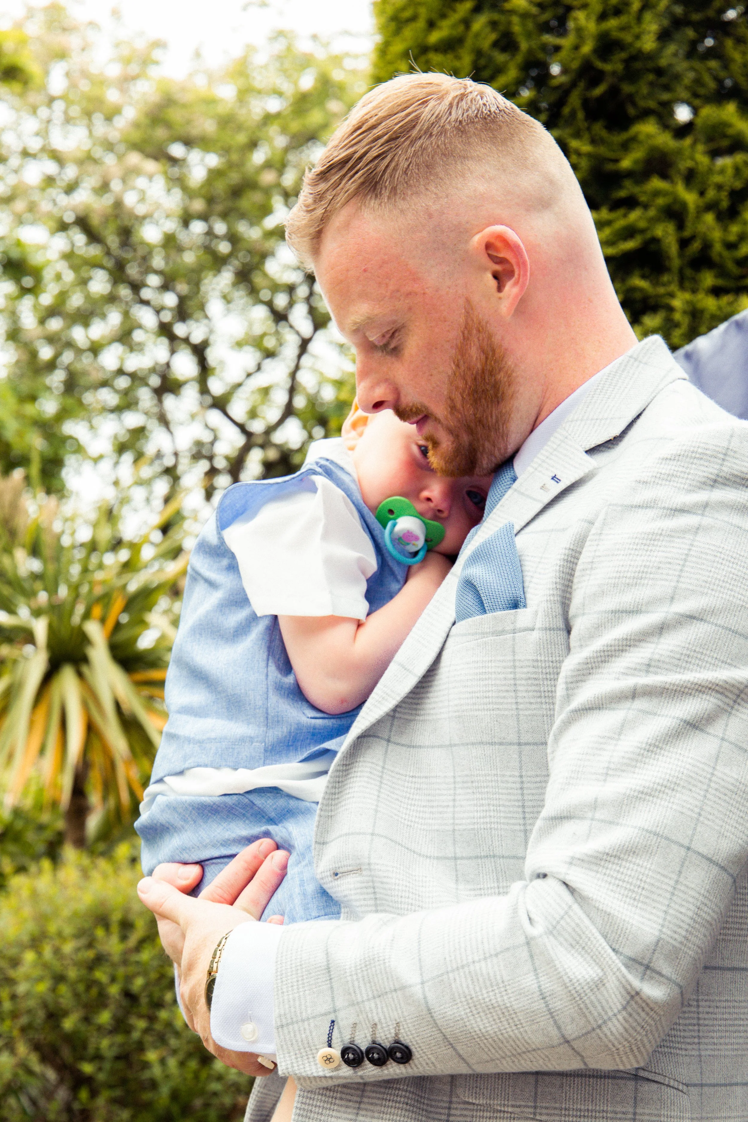 Man in light gray suit holding a sleeping child with a pacifier outdoors.