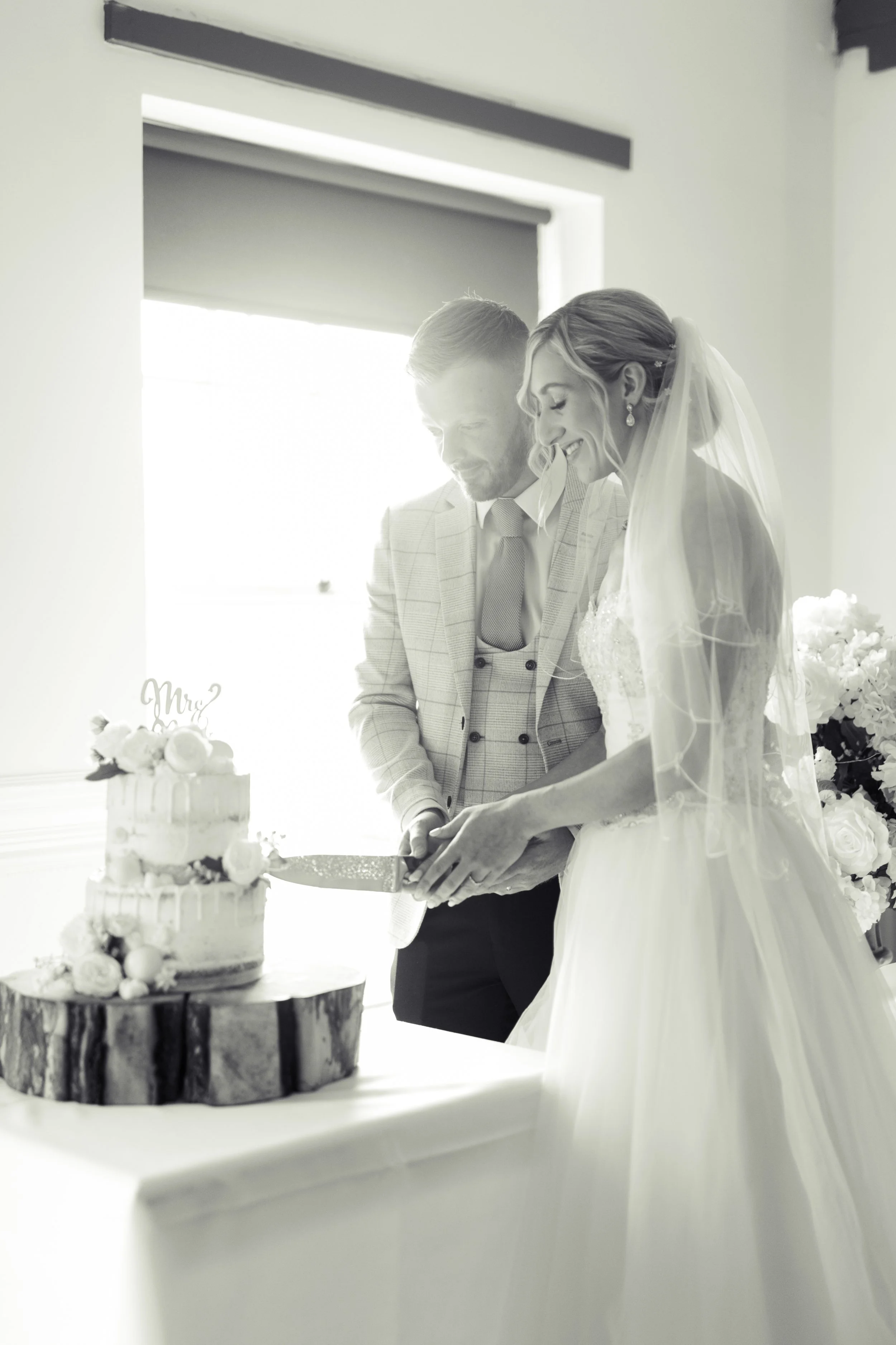 A bride and groom cutting a wedding cake together, with the bride in a wedding dress and veil, and the groom in a suit, in a bright indoor setting.