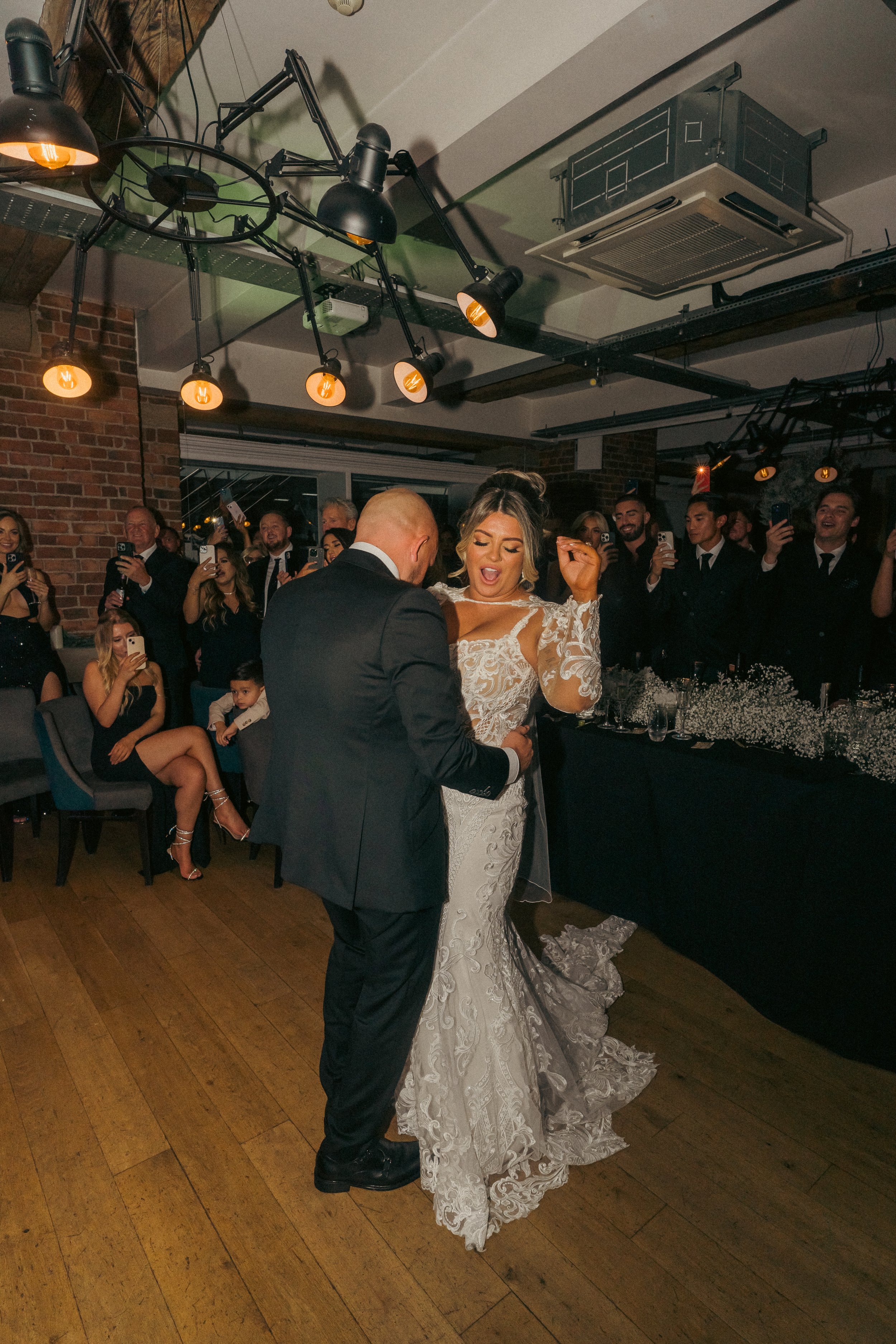 A bride and groom dancing at their wedding reception, surrounded by guests taking photos and videos, in a decorated indoor venue with brick walls and wooden floors.