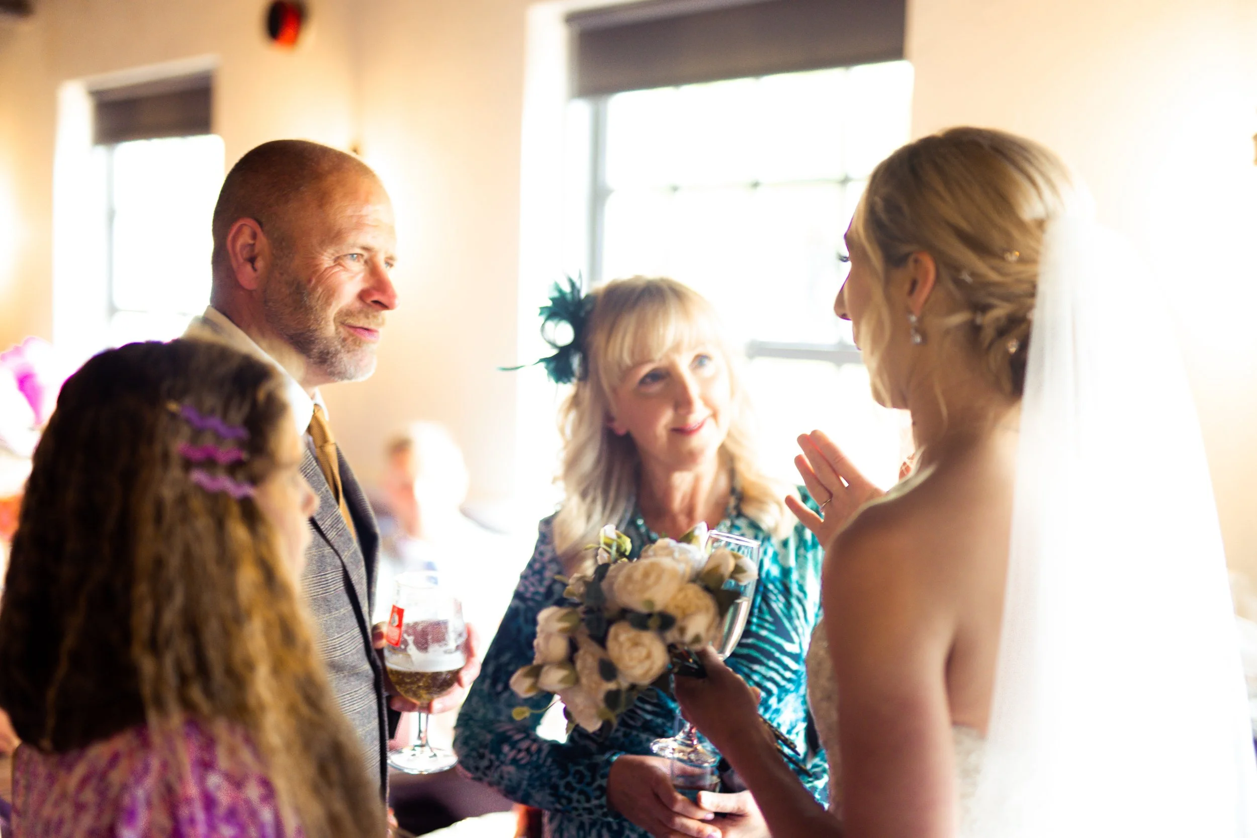 A bride in a wedding dress with a veil talking to guests at her wedding, including an older woman holding a bouquet and a man holding a glass of beer, inside a well-lit room.