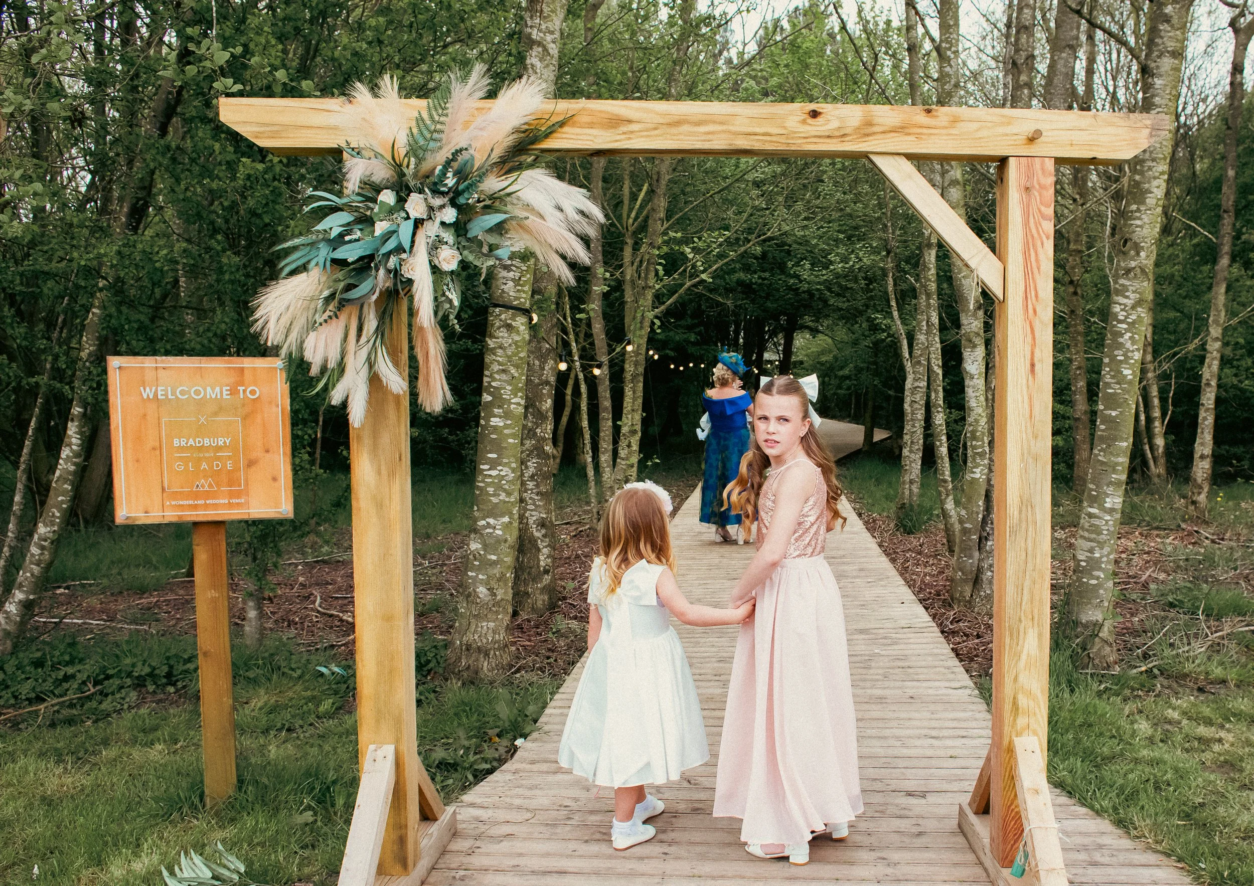 Two young girls dressed in formal dresses holding hands under a wooden wedding arch decorated with white and green pampas grass and flowers, walking down a wooded outdoor path during daytime.