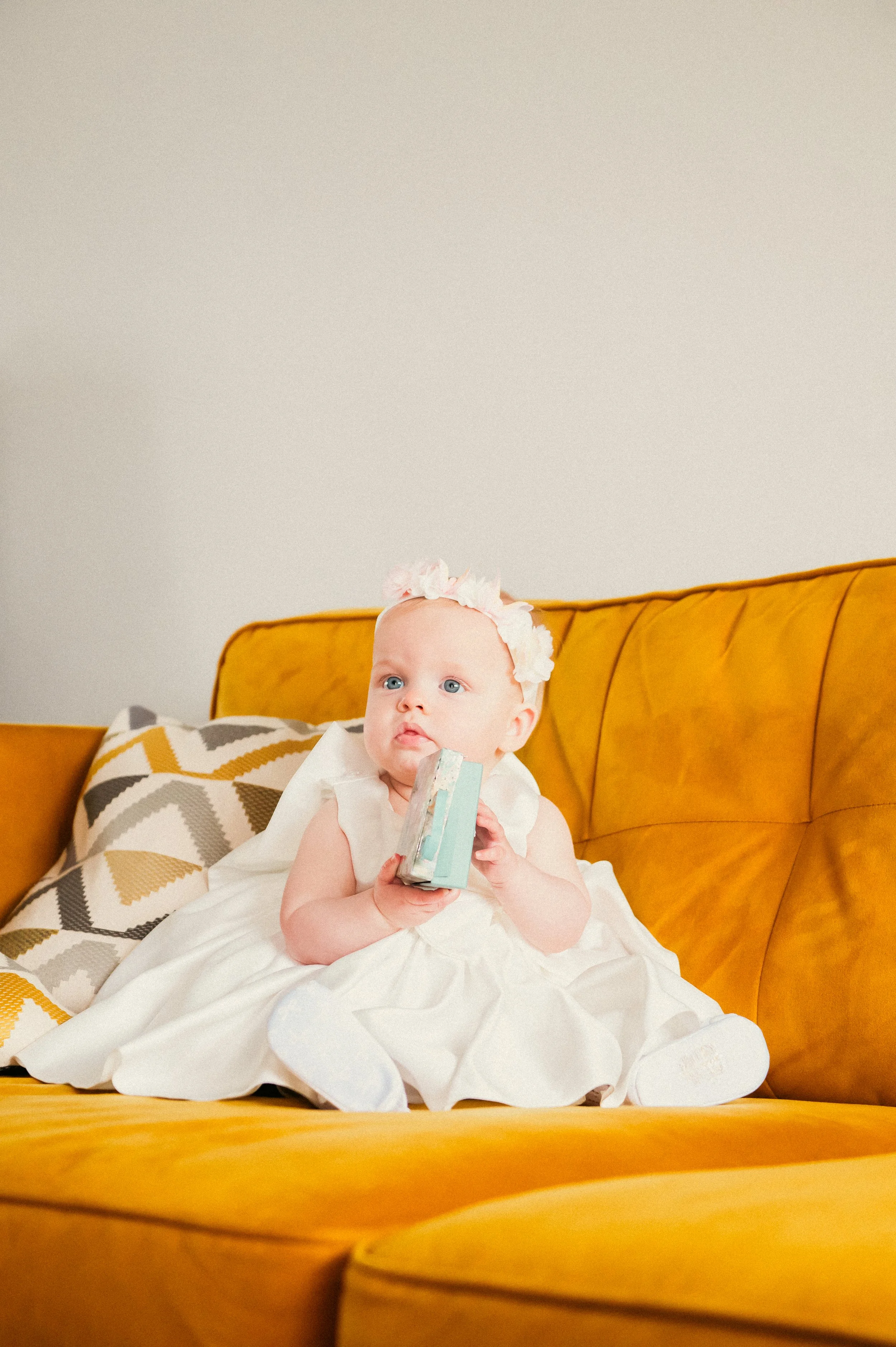 A baby girl with a flower headband sitting on a yellow couch holding a block of bath soap.