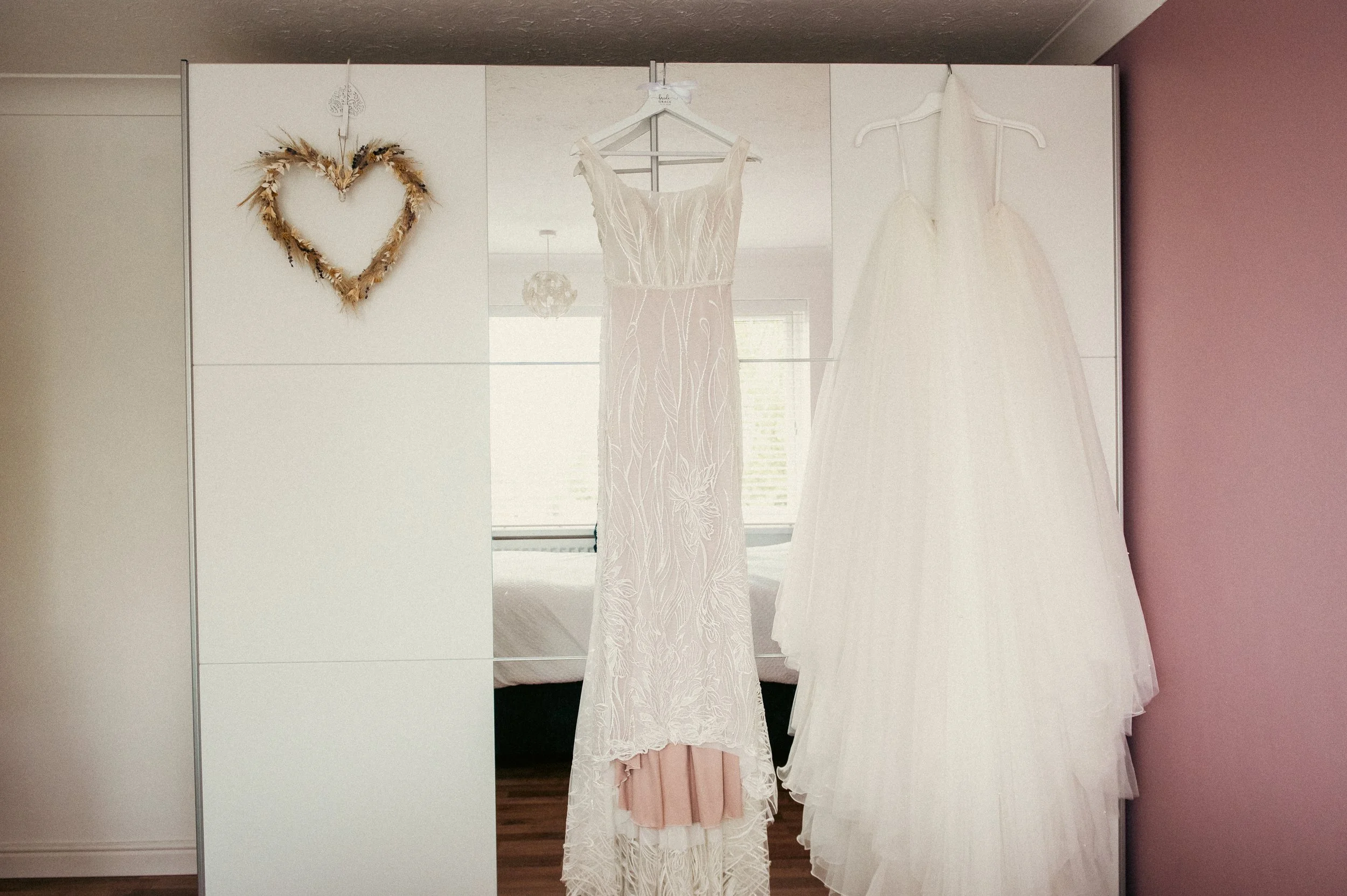 Two wedding dresses hanging on hangers and a heart-shaped wreath on a mirrored closet door in a bedroom.