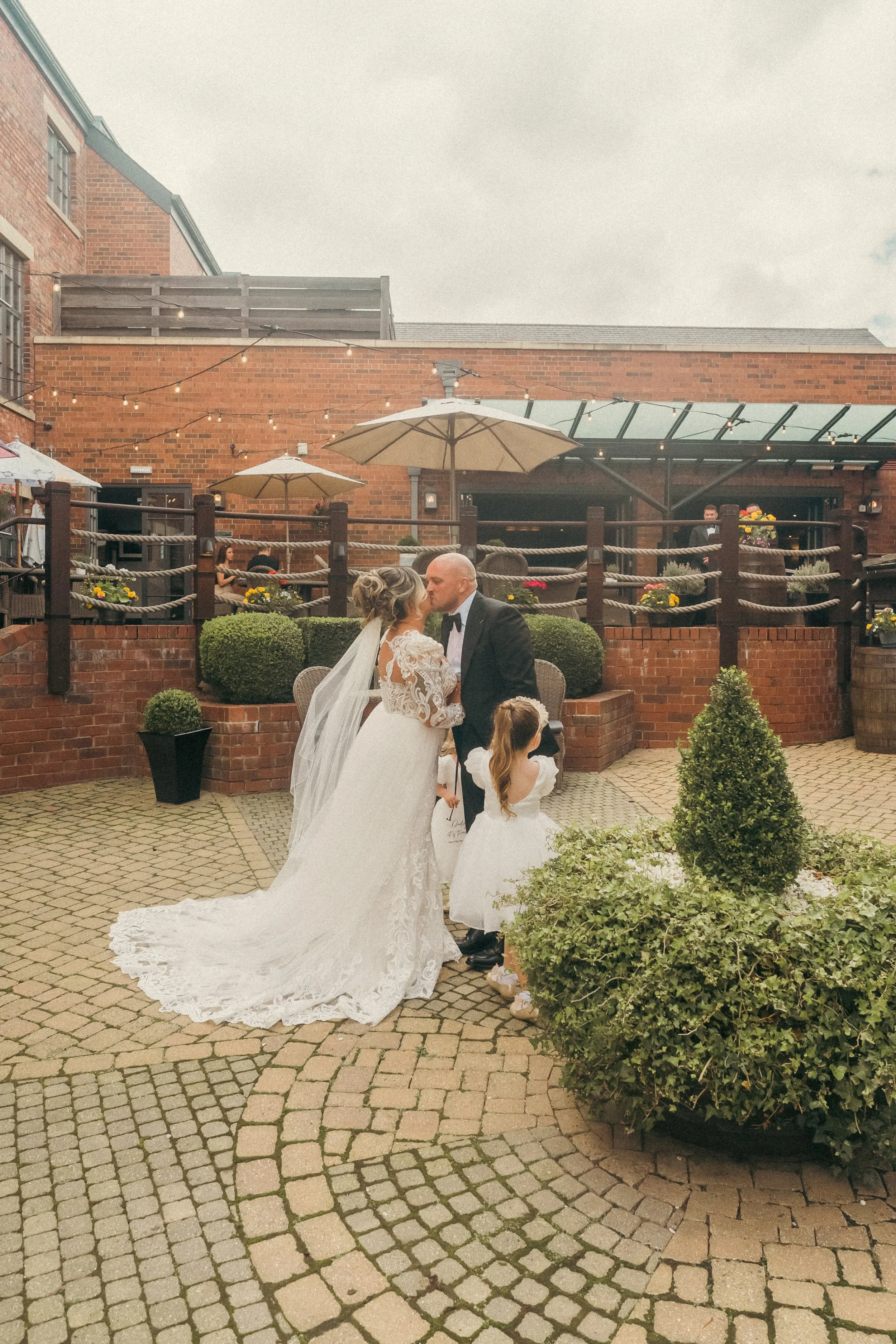 A bride and groom kiss at an outdoor wedding ceremony, with a girl in a white dress watching nearby.