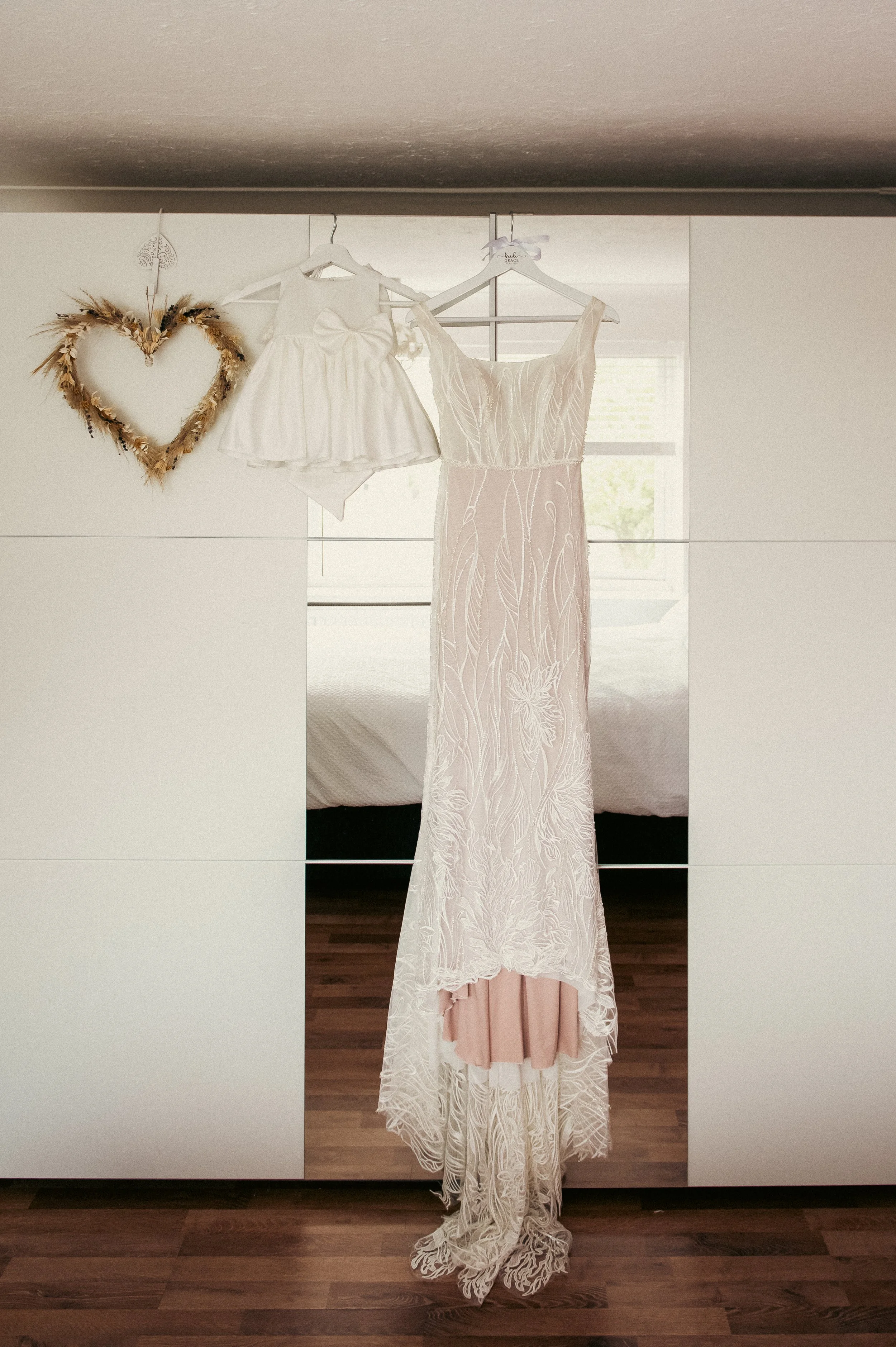 A wedding dress hangs on a hanger in front of a mirror. Two smaller garments, possibly for a flower girl and a ring bearer, hang beside it. A heart-shaped wreath decorates the wall nearby.