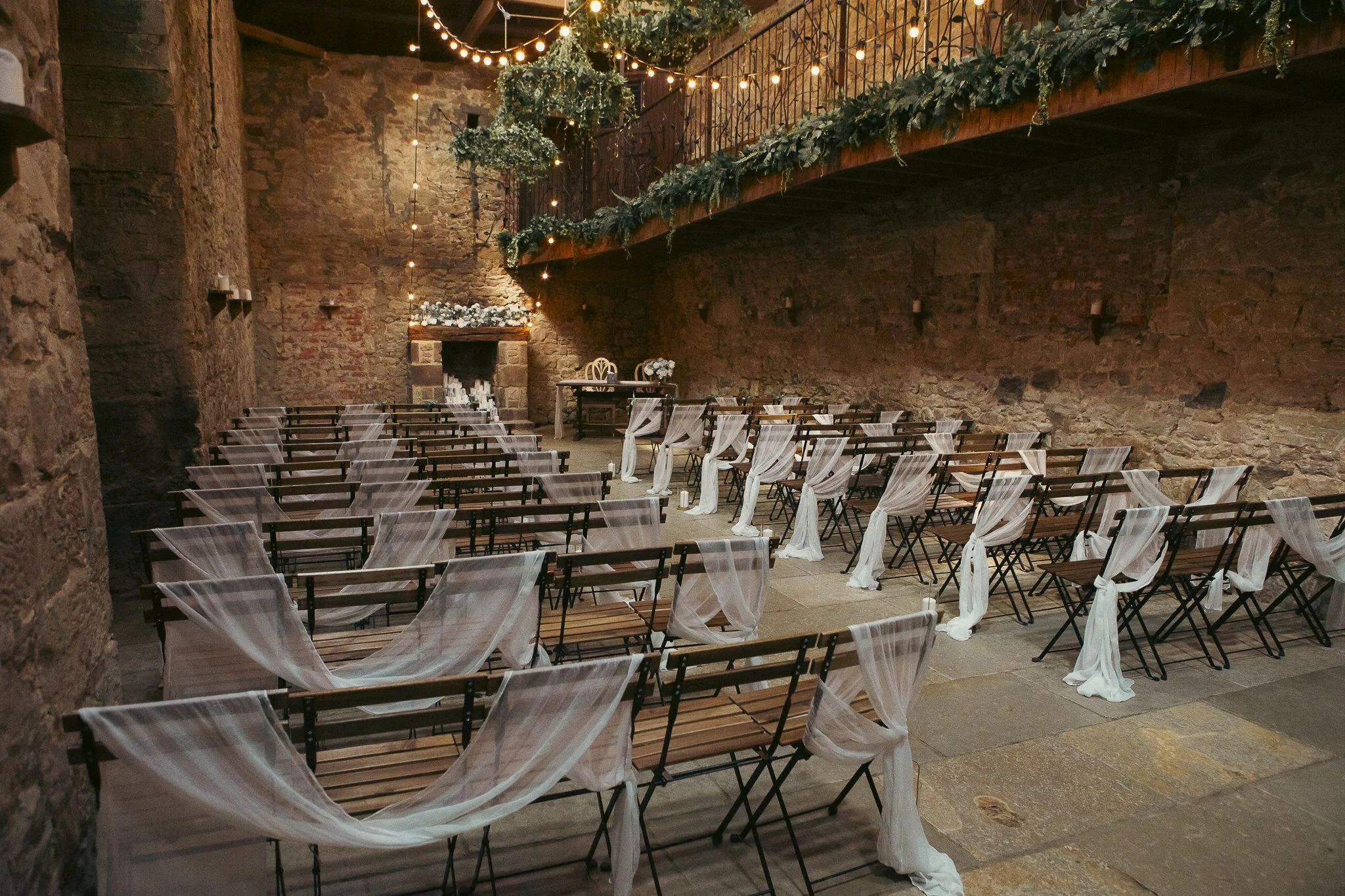 Indoor wedding ceremony setup in a rustic brick hall with chairs decorated with white tulle, a fireplace at the back, and string lights hanging from the ceiling.