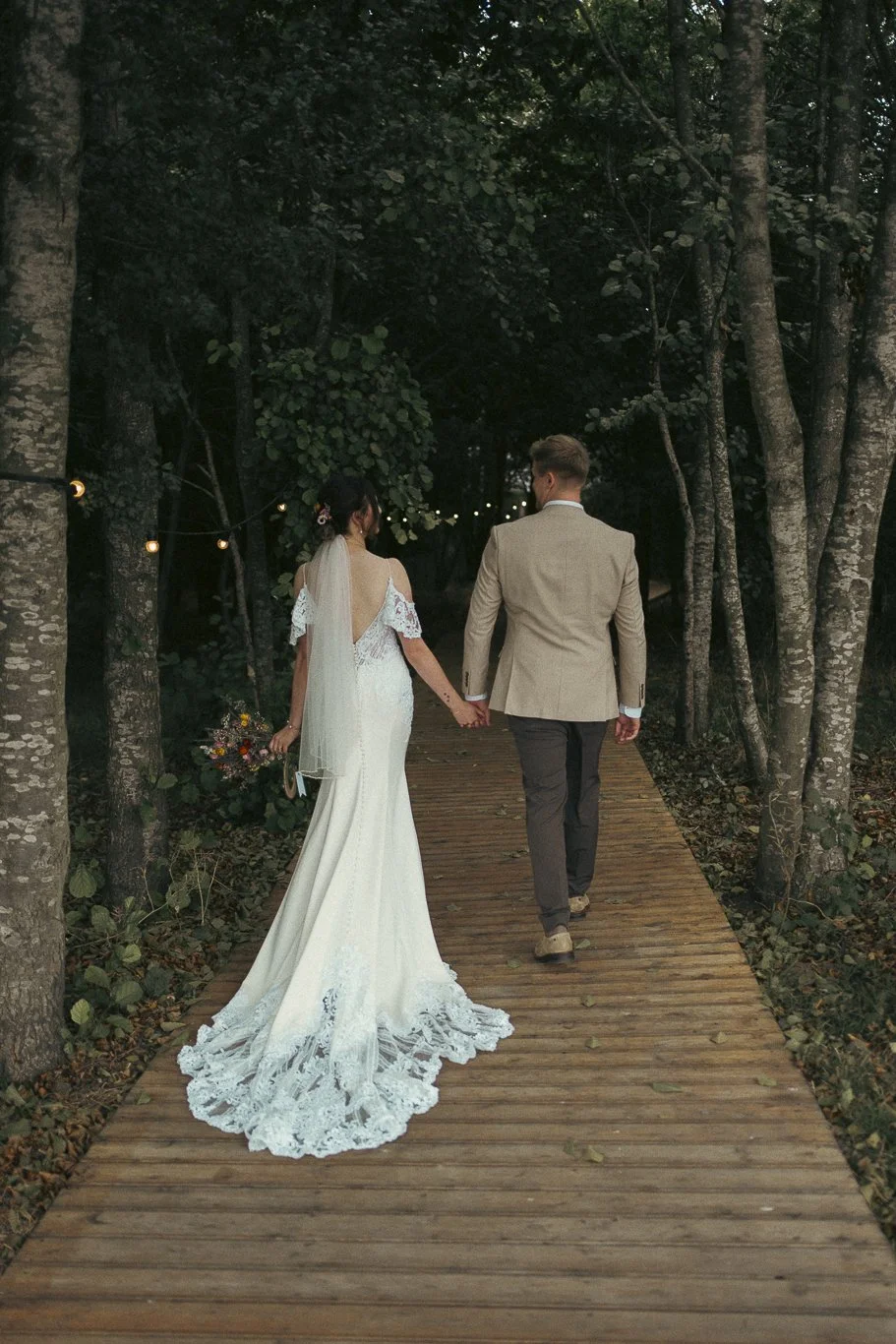 Bride and groom walking hand in hand on a wooden path through a forested area during a wedding, with string lights overhead.