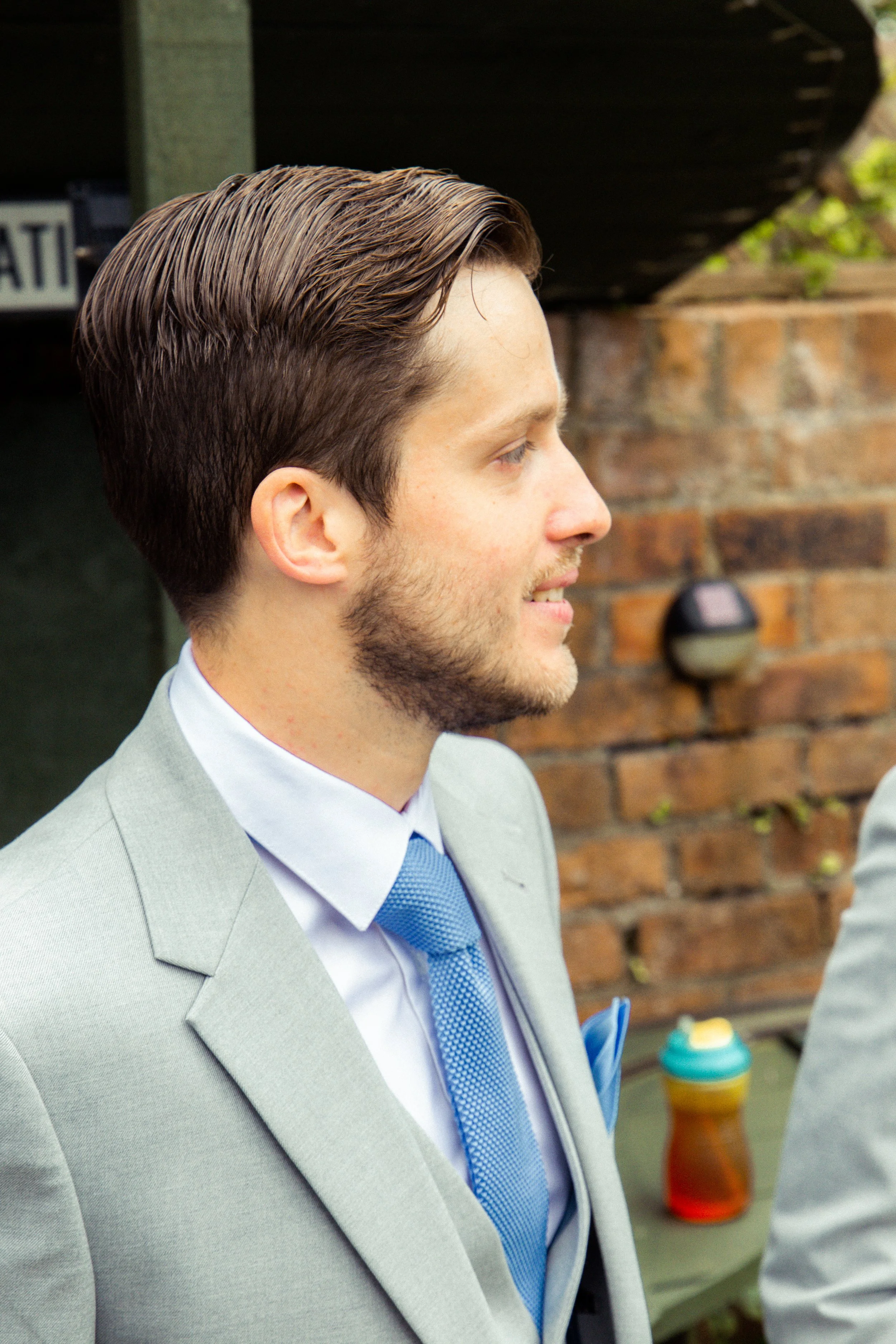Side profile of a man in a grey suit with a blue tie and a light blue shirt, standing outdoors in front of a brick wall and a wooden structure.