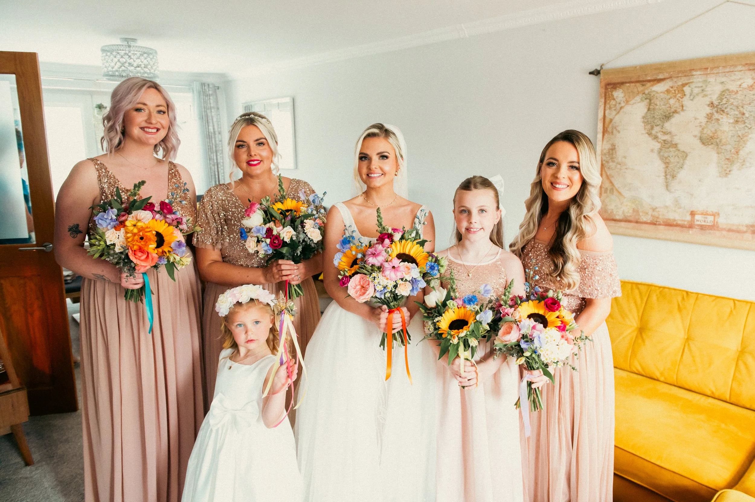 A bride and five women, with two young girls, standing indoors holding colorful bouquets, in a room with a world map on the wall and a yellow sofa.