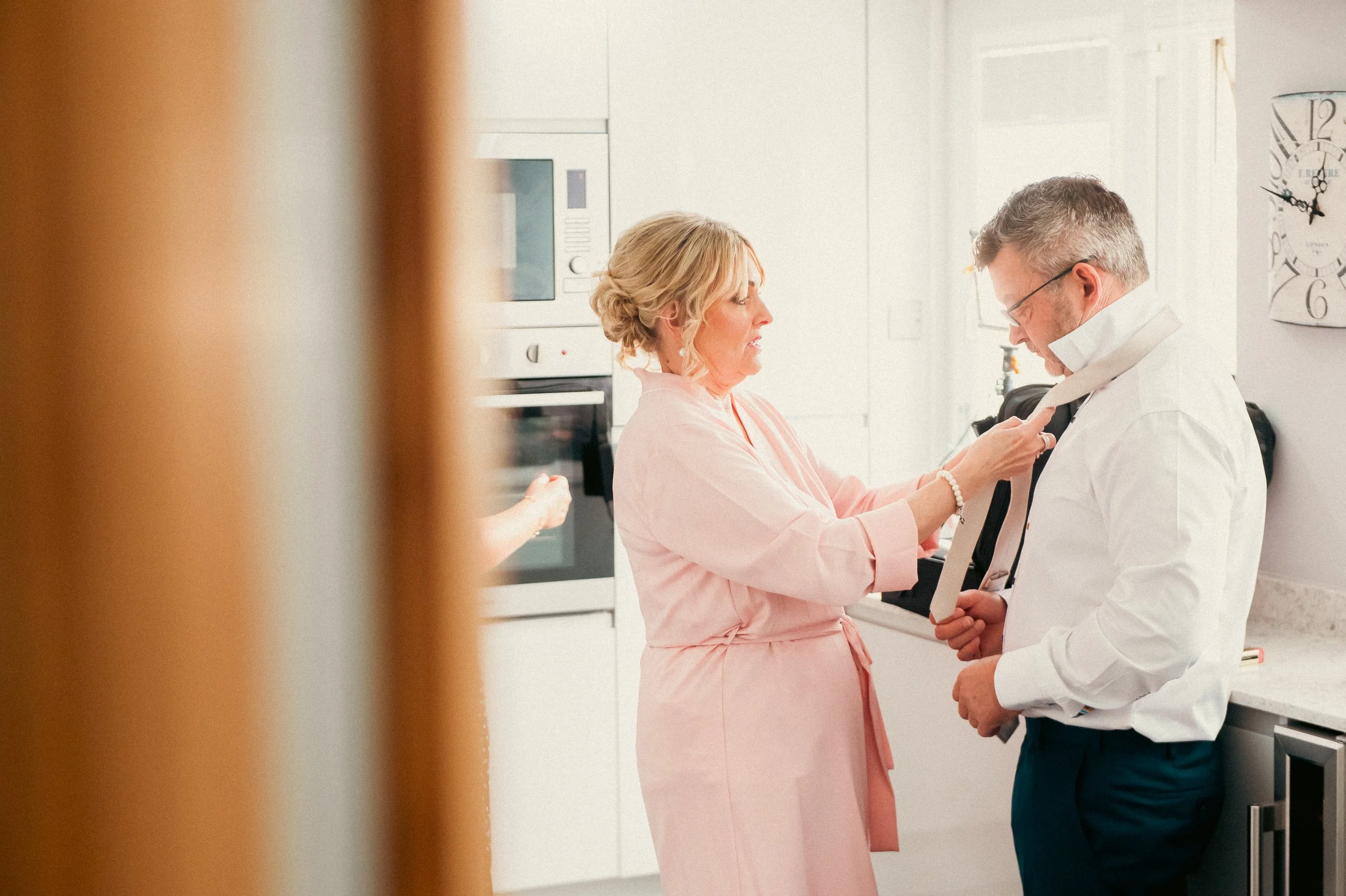A woman in a pink robe helping a man in a white shirt with a tie in a kitchen.