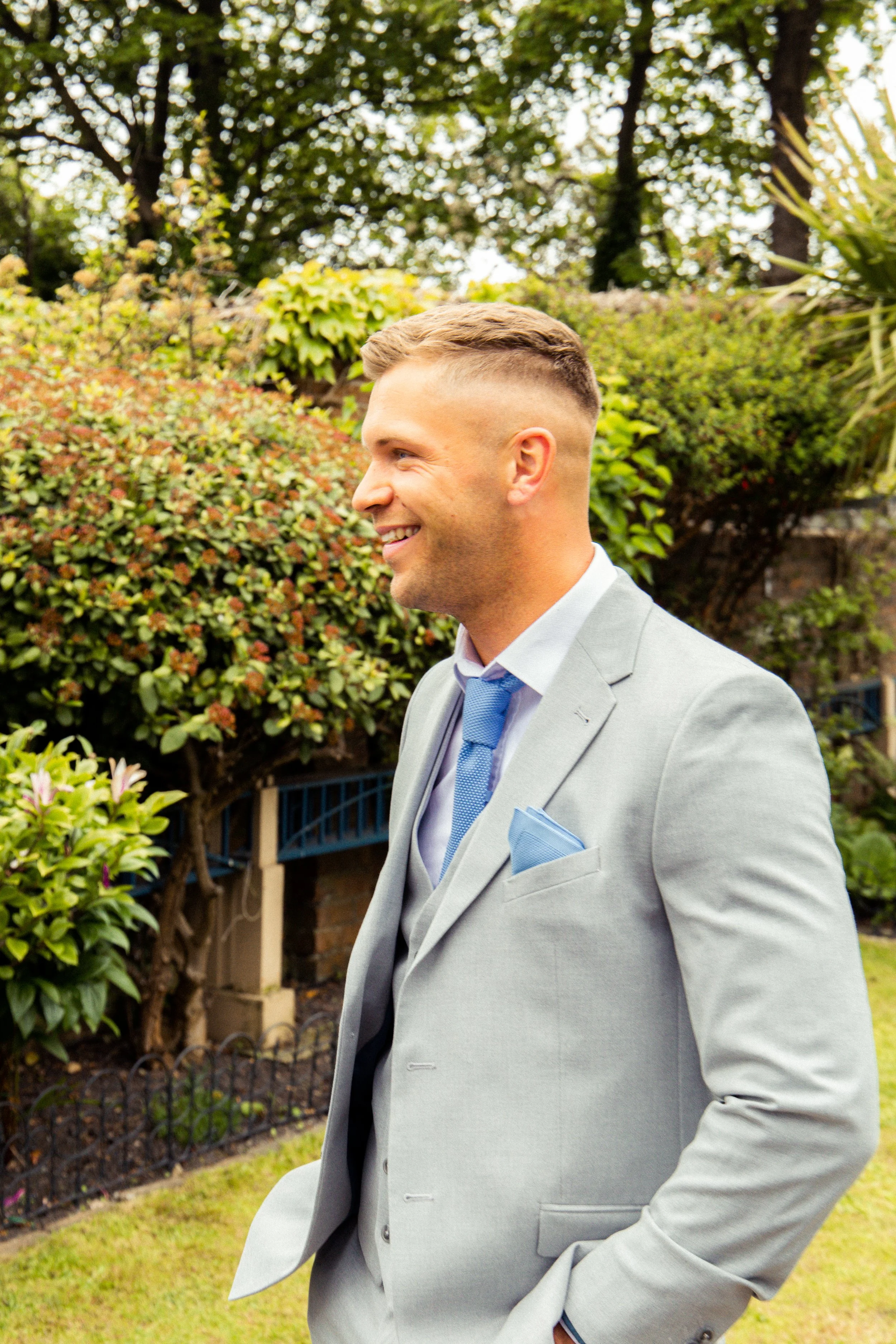 A man in a light gray suit with a blue tie and pocket square, smiling in a garden with bushes and trees in the background.