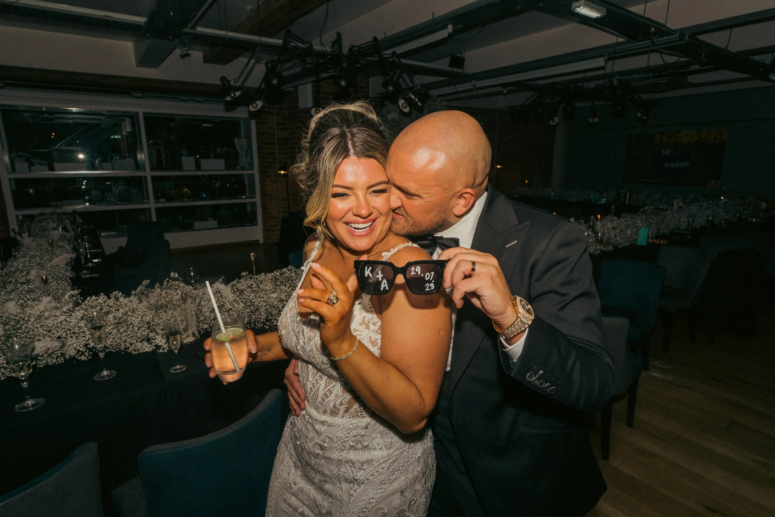 A joyful couple at a wedding reception, with the woman smiling and the man kissing her cheek, holding wedding sunglasses with initials and date, in a decorated venue.