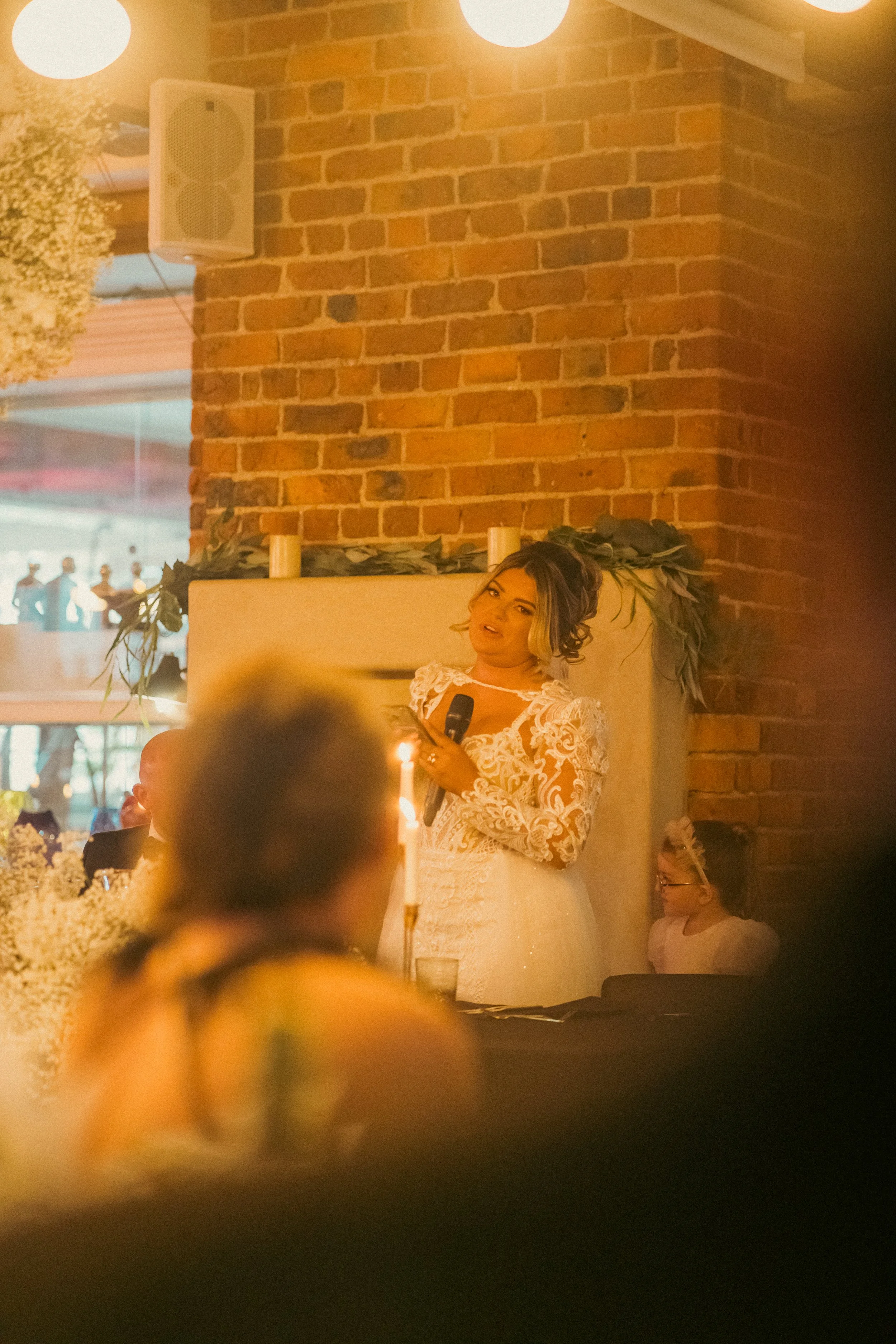 Woman in a white lace dress holding a microphone, speaking at a celebration, with a candle on the table in front of her, brick wall in the background, and children sitting nearby.