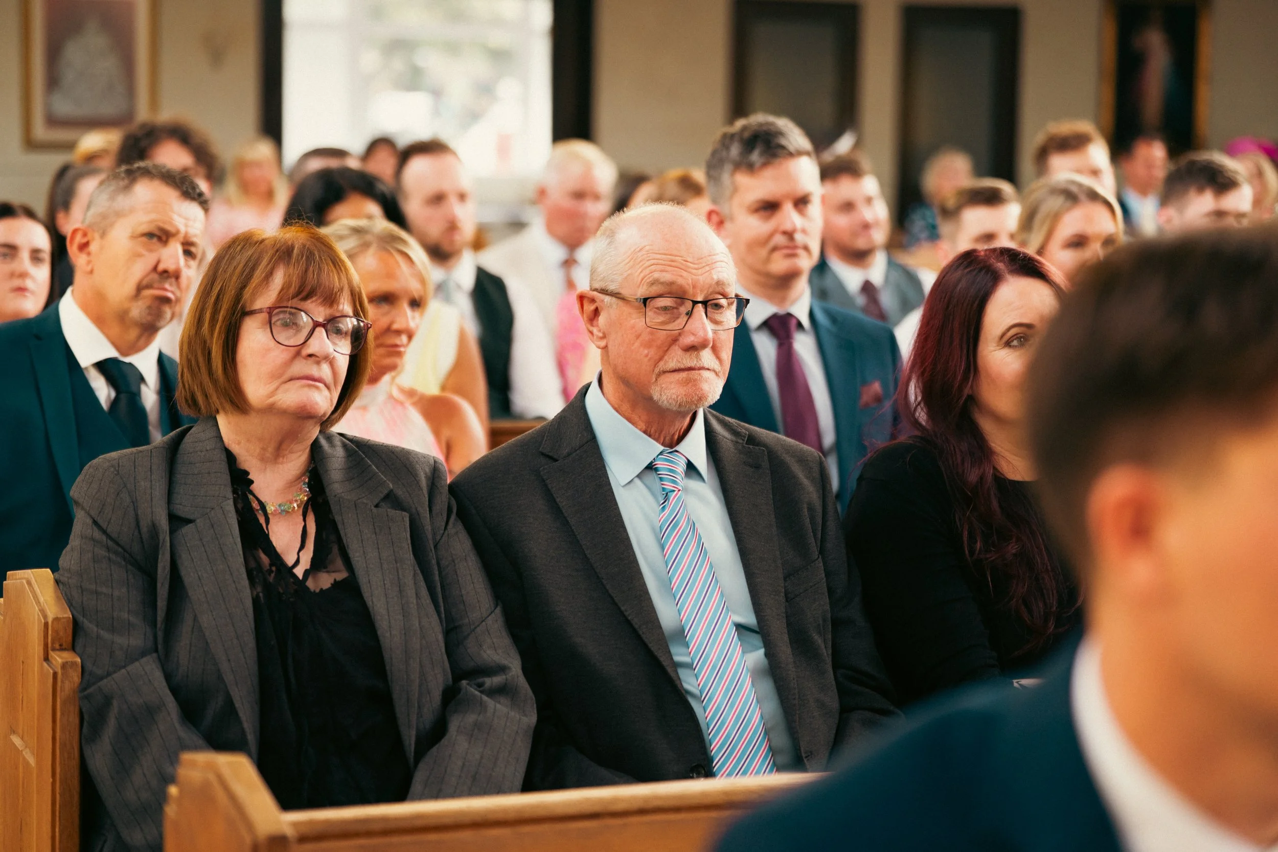 People attending a formal event in a church or hall, dressed in suits and dresses, sitting on wooden pews, with expressions of focus and seriousness.
