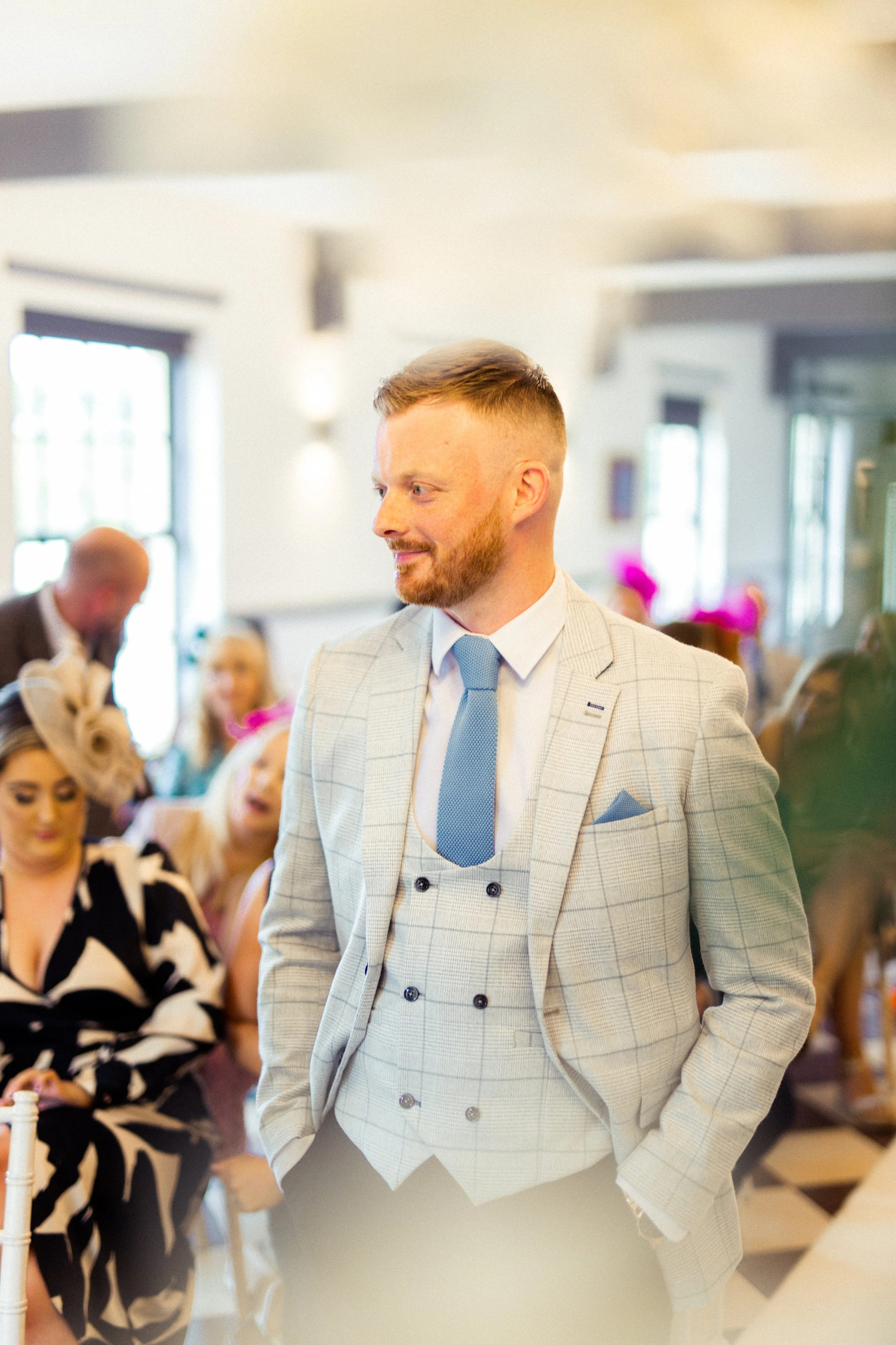 Man in a light checkered suit with a light blue tie, standing with hands in pockets at social event with people in the background.