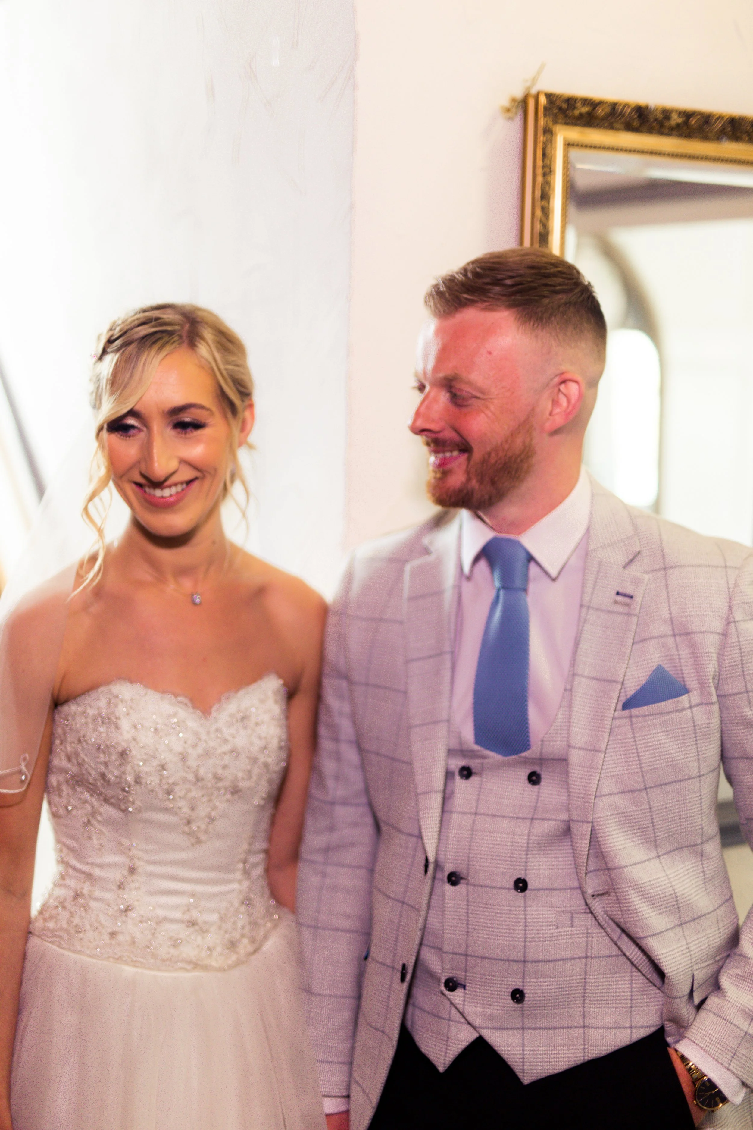 A bride in a white wedding gown and a groom in a light plaid suit with a blue tie, smiling and looking at each other during their wedding celebration.