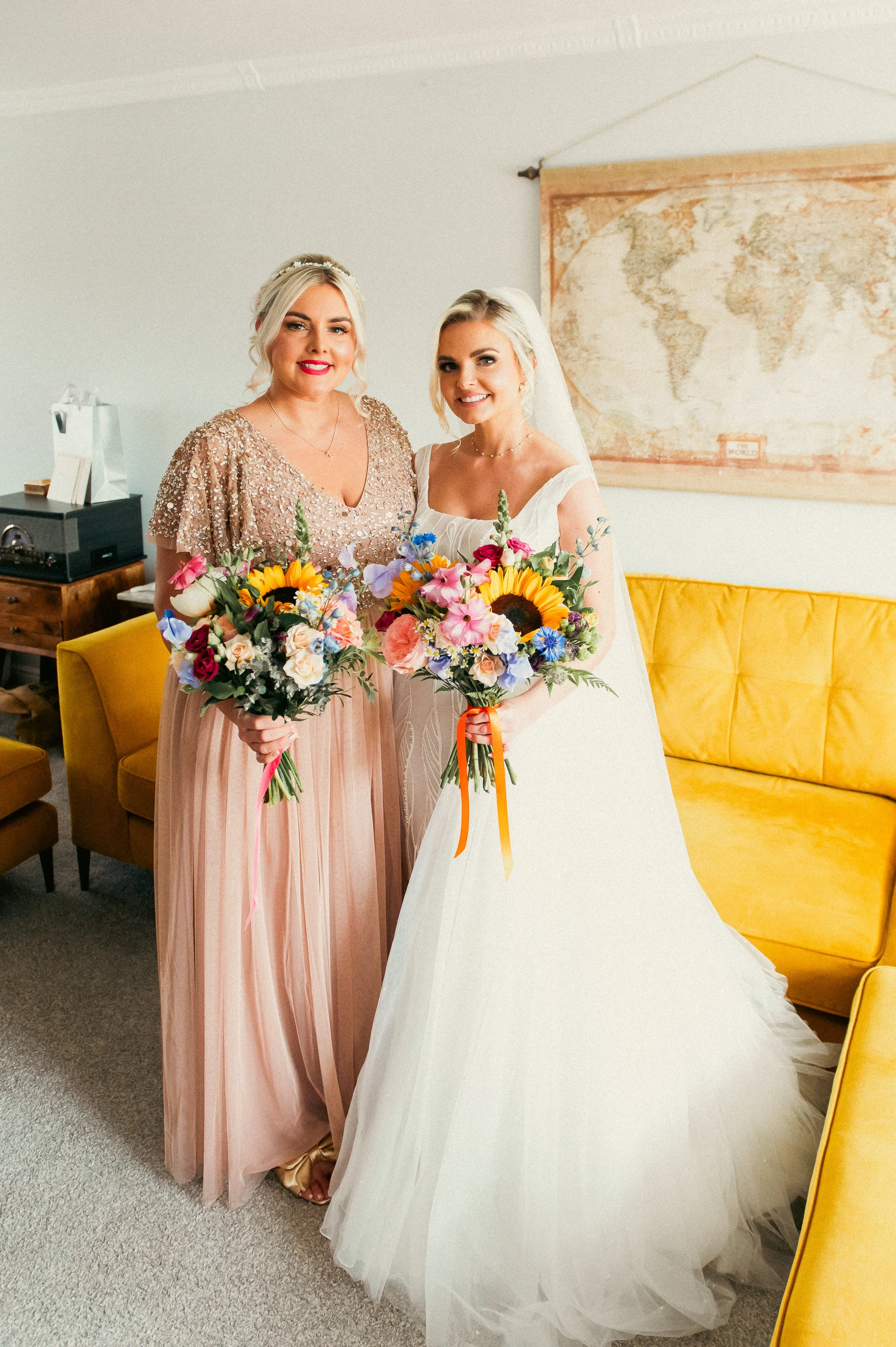 A bride in a white wedding dress and a woman in a pink dress holding colorful bouquets of flowers, standing indoors in front of a yellow couch and a world map on the wall.