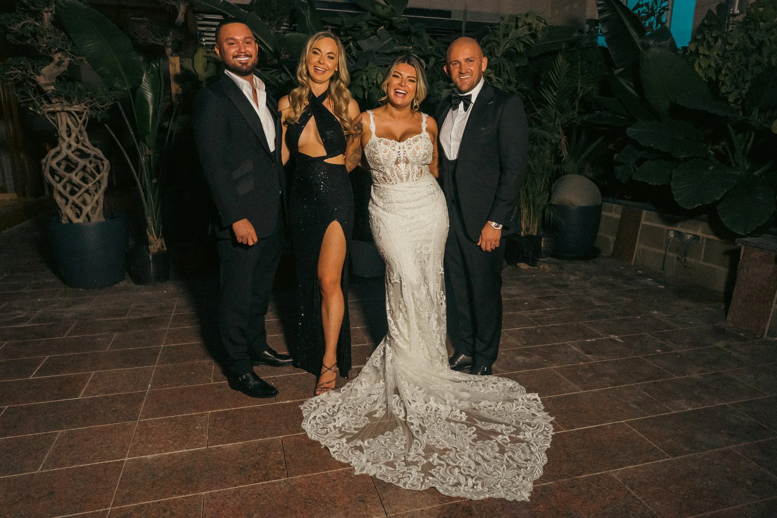 Group of four people dressed formally, standing on a tiled floor with greenery behind them at night. One woman wears a white lace wedding dress, and a woman next to her wears a black gown with a thigh-high slit. Two men are in black suits, one with a