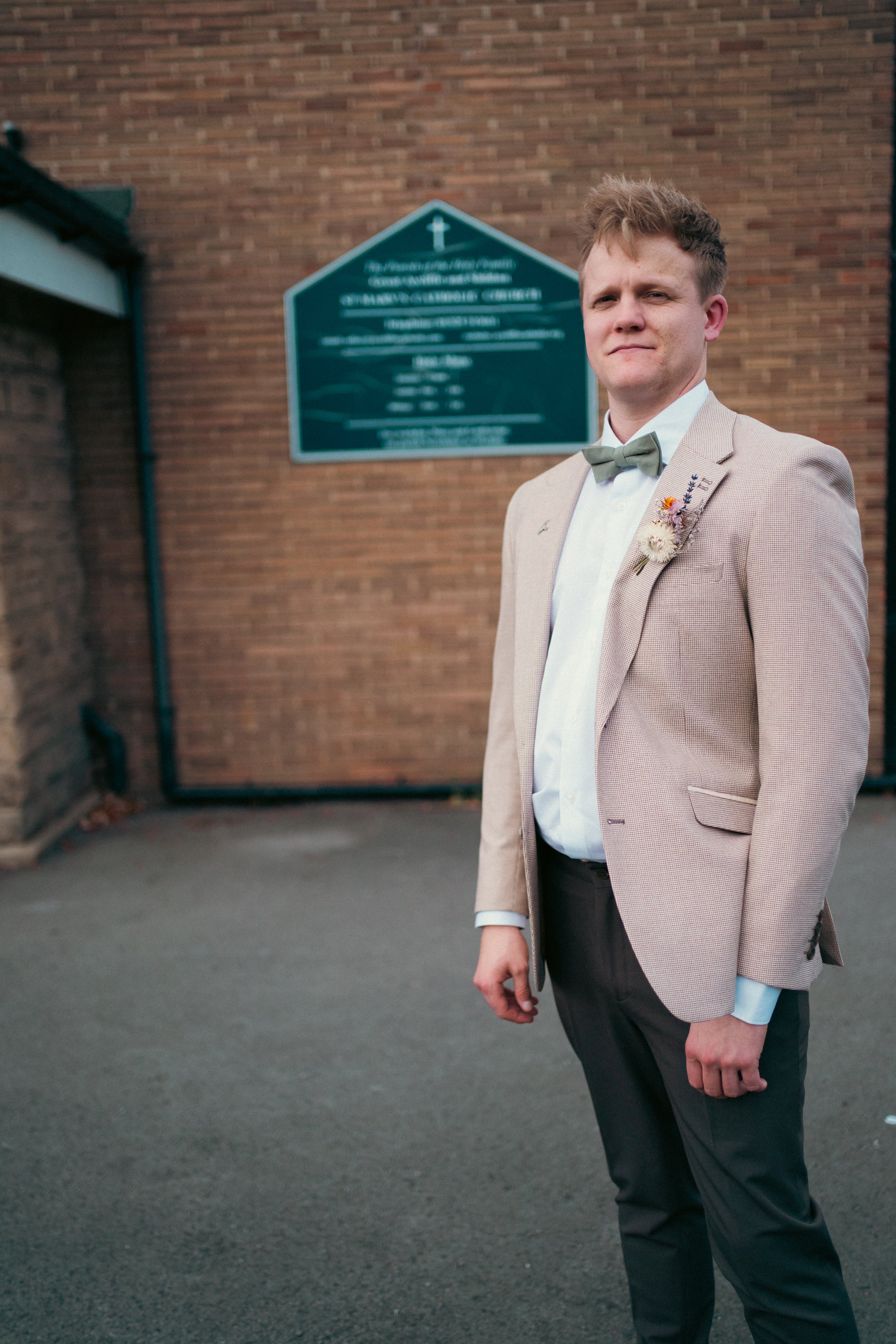 A young man in a beige suit with a gray bow tie standing outdoors in front of a brick wall and a church sign.