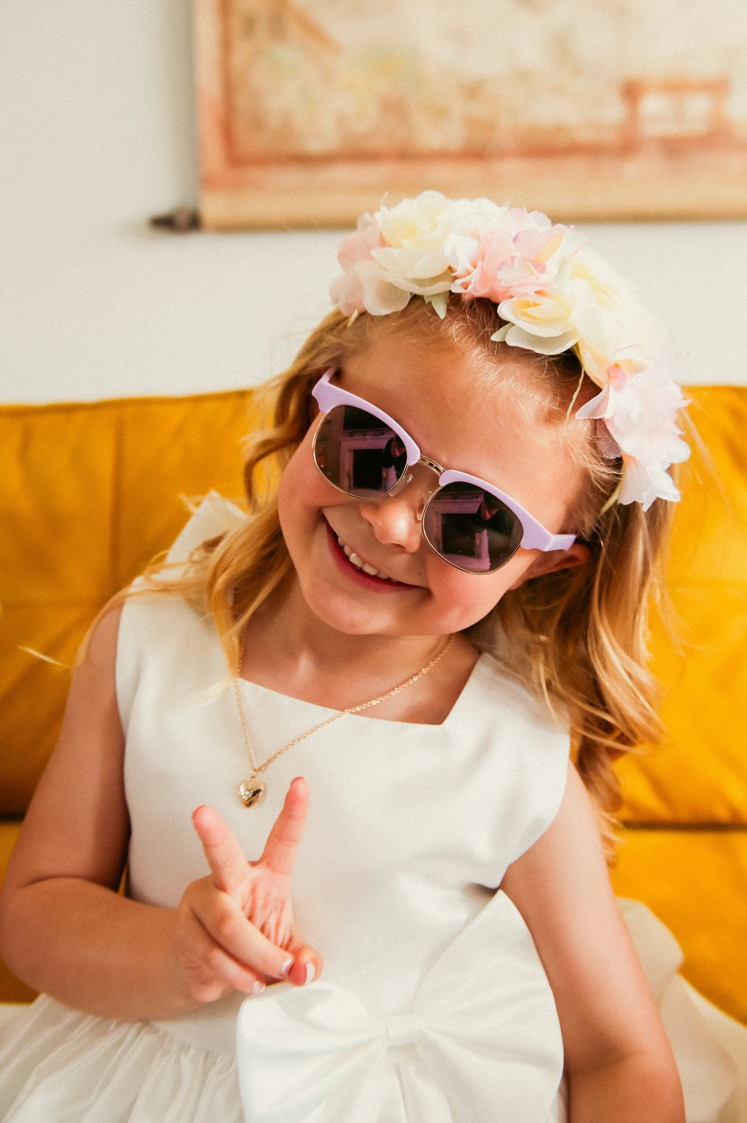 A young girl with blonde hair wearing large sunglasses, a white dress with a bow, a gold necklace with a heart pendant, and a flower crown, sitting on a yellow couch and smiling at the camera.