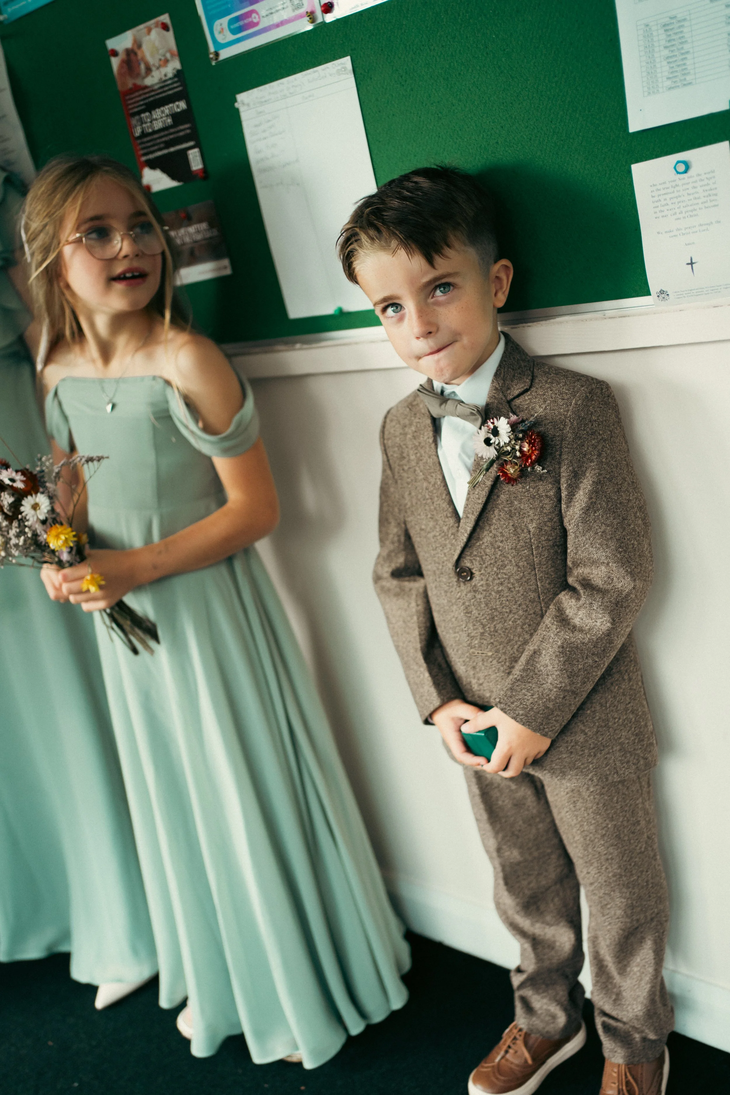 A boy in a brown suit with a boutonniere and a girl in a light teal dress with a boutonniere, standing indoors against a green bulletin board.