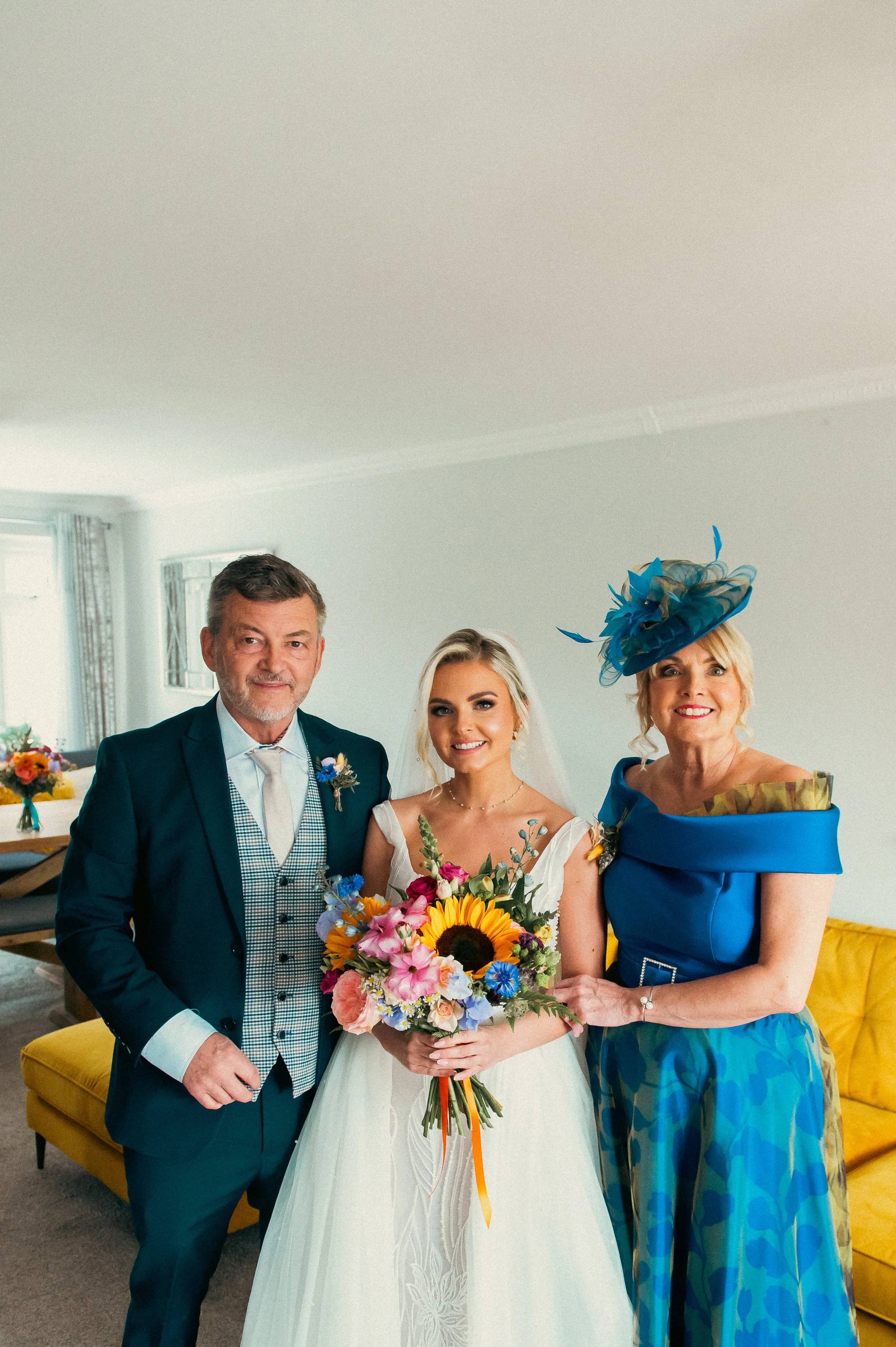 A bride standing with a man and a woman in a brightly lit living room. The bride is holding a colorful bouquet with a sunflower. The woman on the right is wearing a vibrant blue dress with a matching hat, and the man is dressed in a suit with a blue 