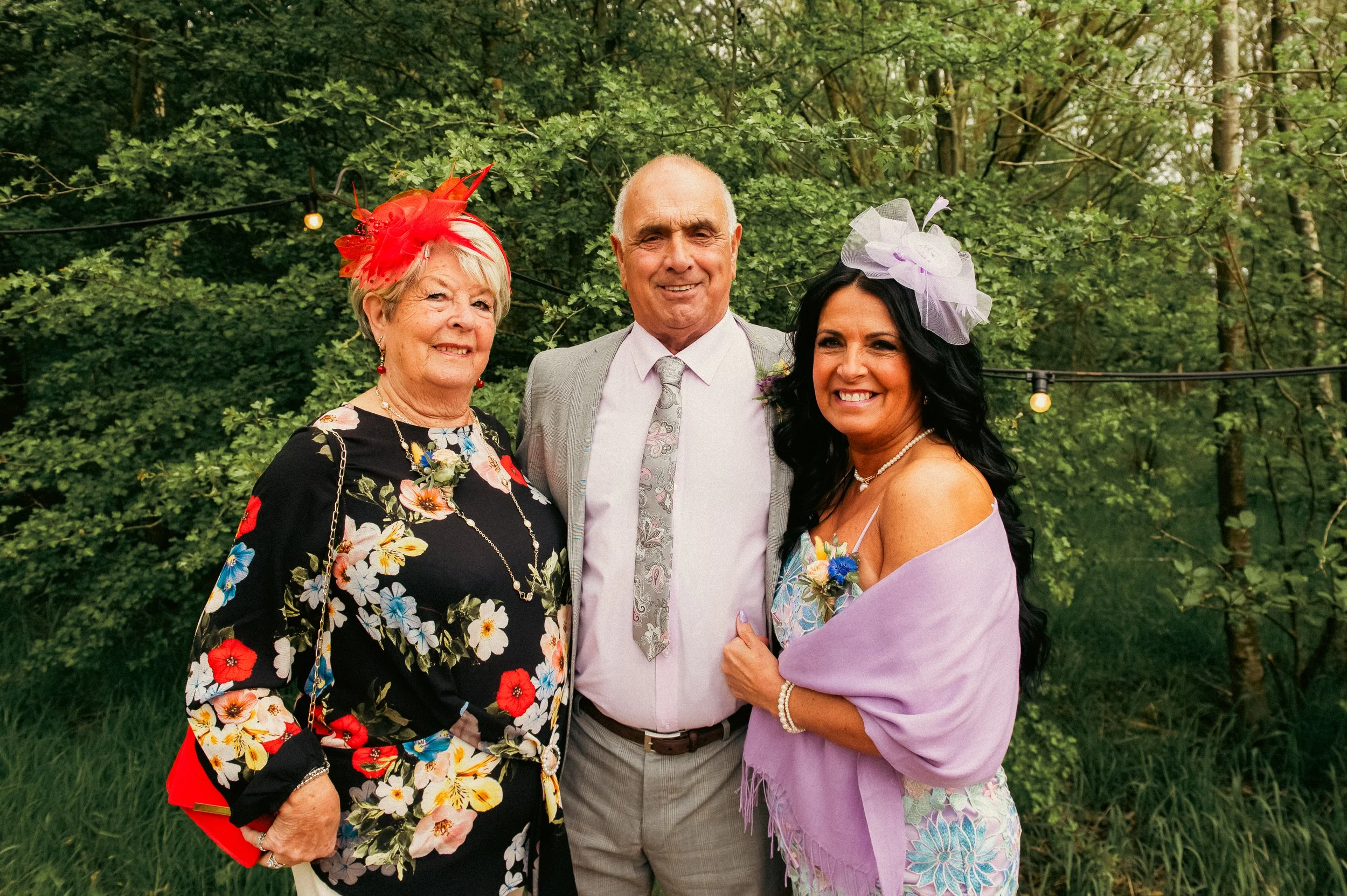 Three people, two women and one man, smiling and dressed formally outdoors at a garden wedding. The woman on the left wears a black floral dress with a red fascinator. The man in the middle wears a light grey suit with a white shirt and patterned tie